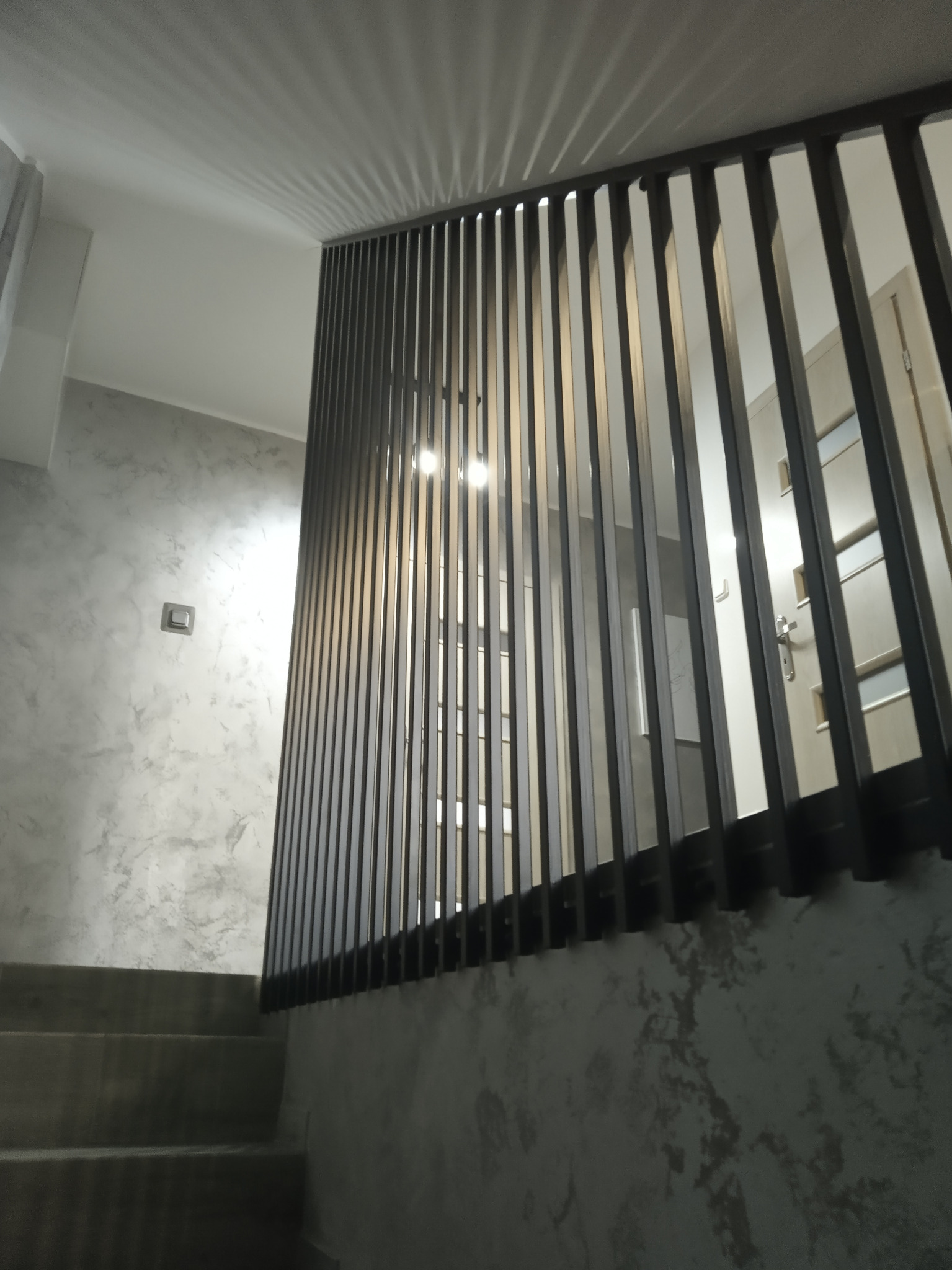 Nowoczesna balustrada schodowa z pionowych, metalowych elementów w kolorze grafitowym, zamontowana przy schodach o betonowej fakturze, widok z dołu, oświetlenie sufitowe.