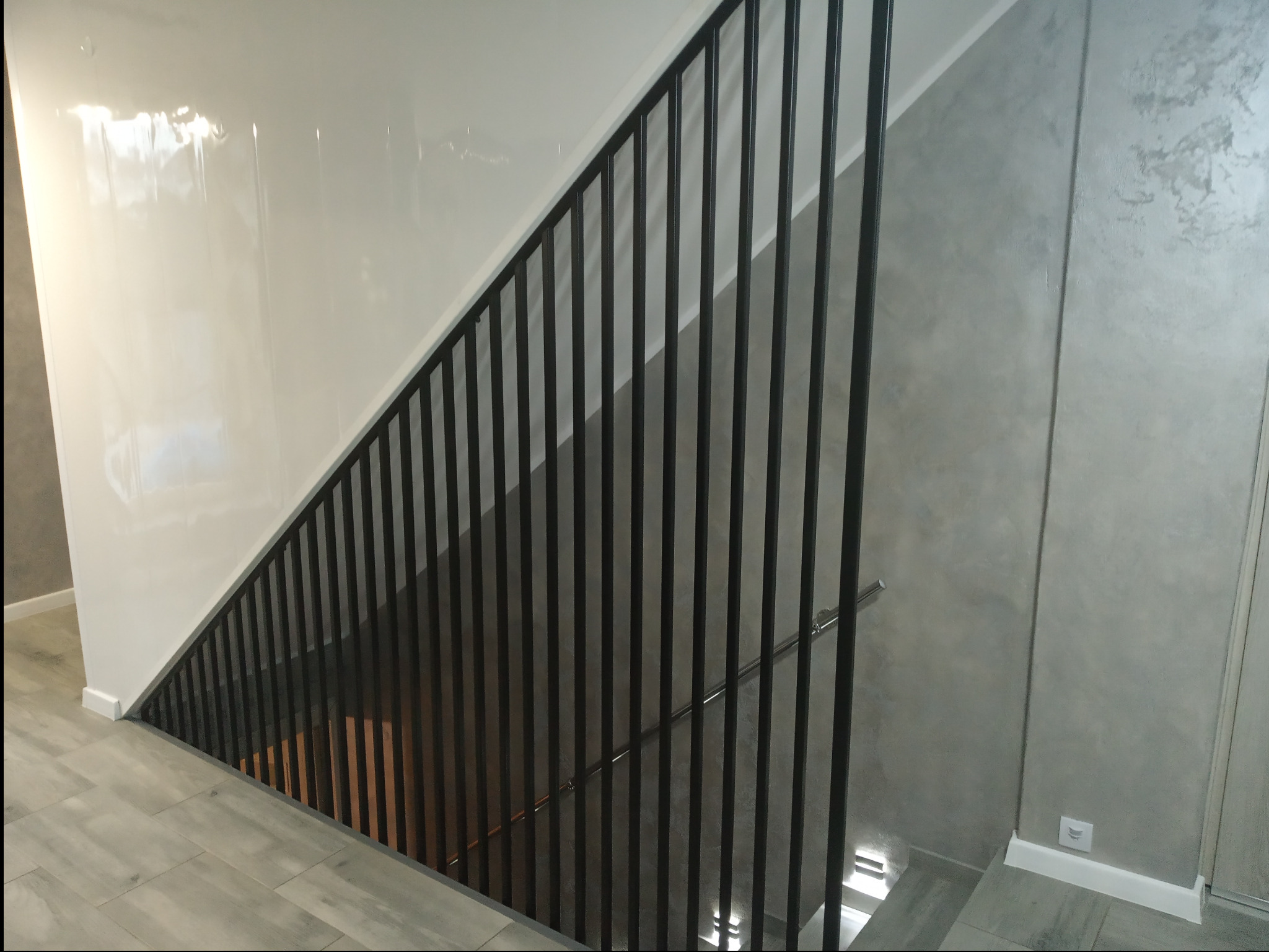 Nowoczesne schody z czarną metalową balustradą, widok z góry na szare płytki podłogowe i białą ścianę z połyskiem.