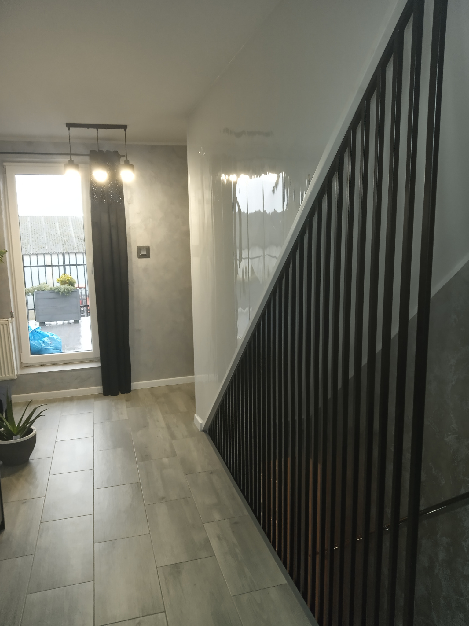 Nowoczesne schody metalowe z czarną balustradą z pionowych prętów, widok z dołu na tle szarej ściany i podłogi z płytek ceramicznych, w tle okno z widokiem na zewnątrz.