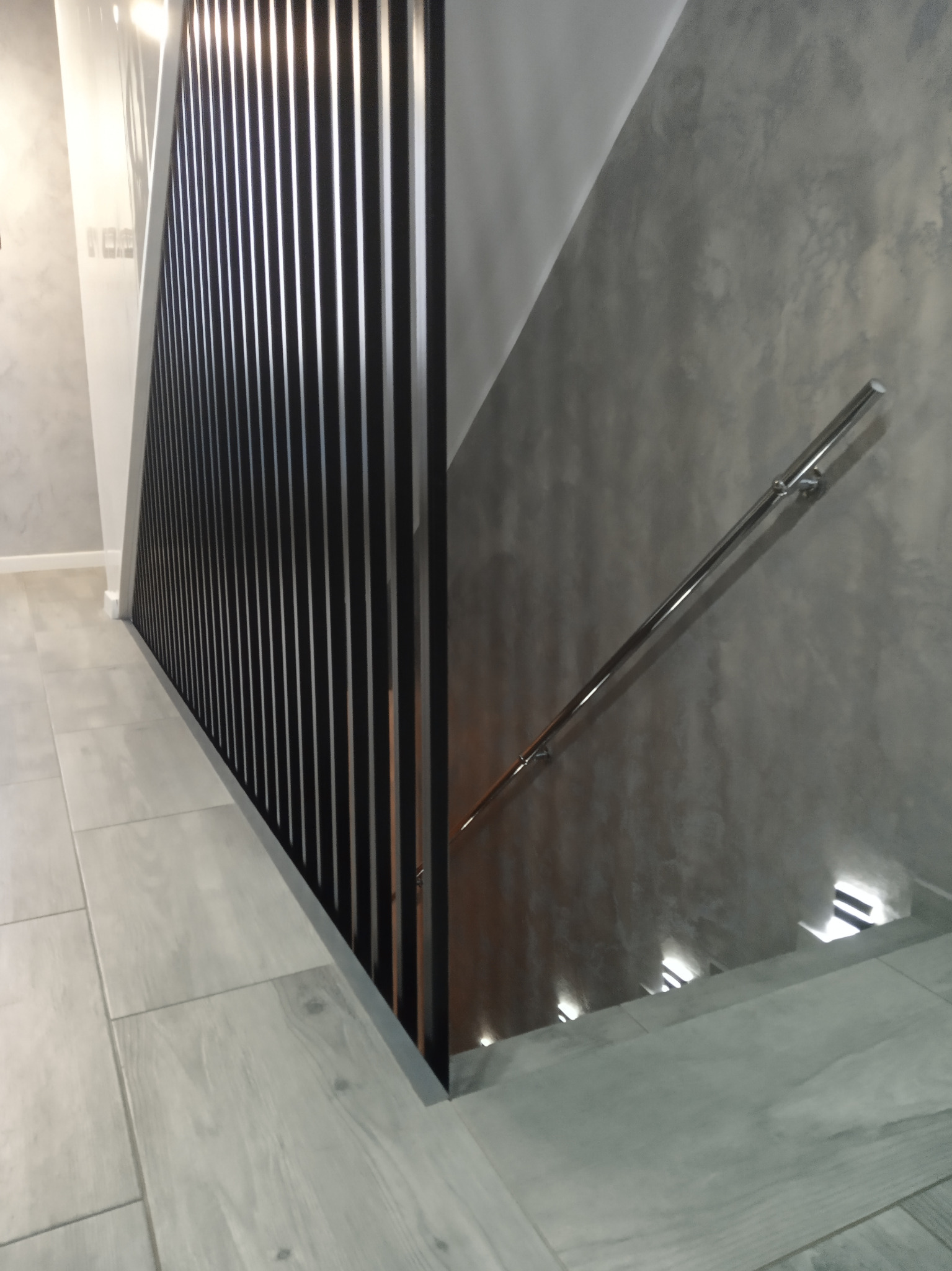 Nowoczesne schody wewnętrzne z czarną, pionową balustradą, szarą ścianą o fakturze betonu i chromowaną poręczą, oświetlone punktowo od dołu.