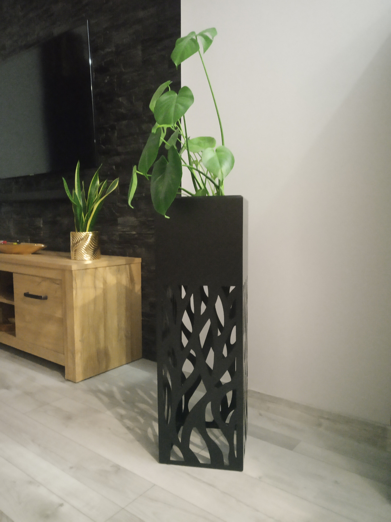 Czarna, metalowa donica z ażurowym wzorem, stojąca obok drewnianej komody z rośliną w złotej osłonce, na tle ściany z imitacji kamienia i jasnej ściany.