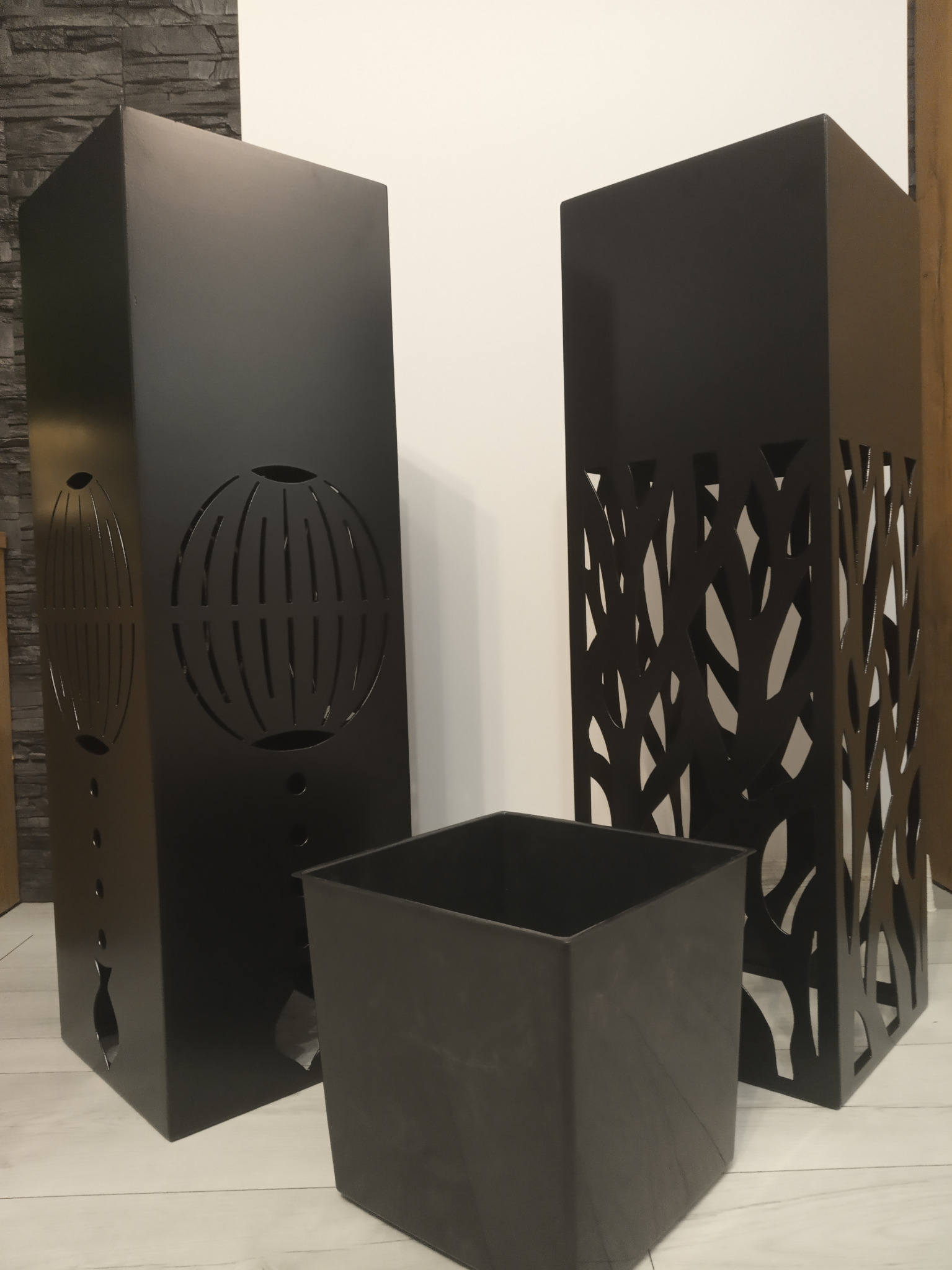 Dwa czarne, metalowe słupki dekoracyjne z wycinanymi wzorami (globus i gałęzie) oraz czarny, kwadratowy pojemnik na podłodze z paneli.