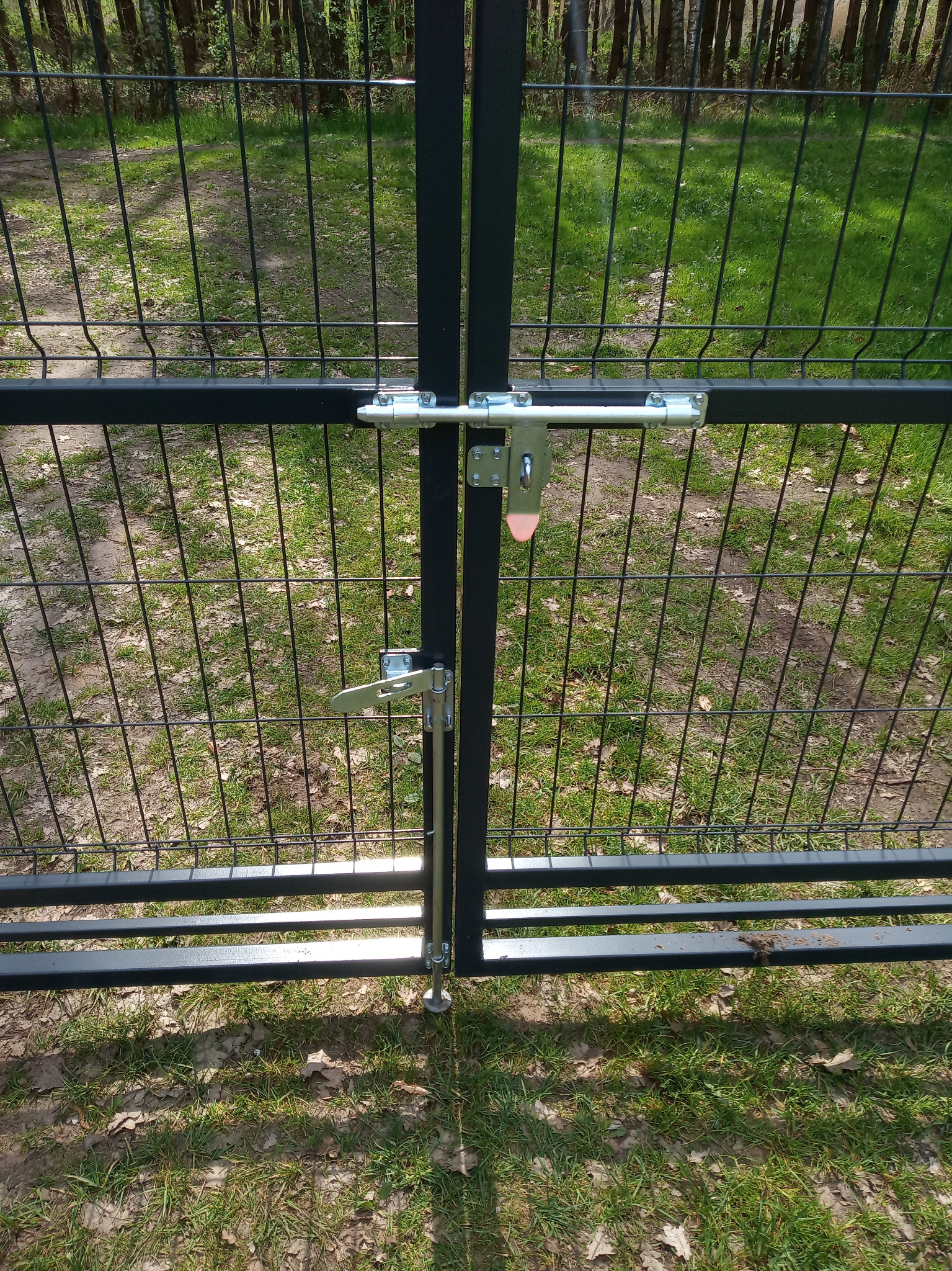 Czarna brama z metalowej siatki z dwoma różnymi typami zamków, widok z bliska na tle zieleni.