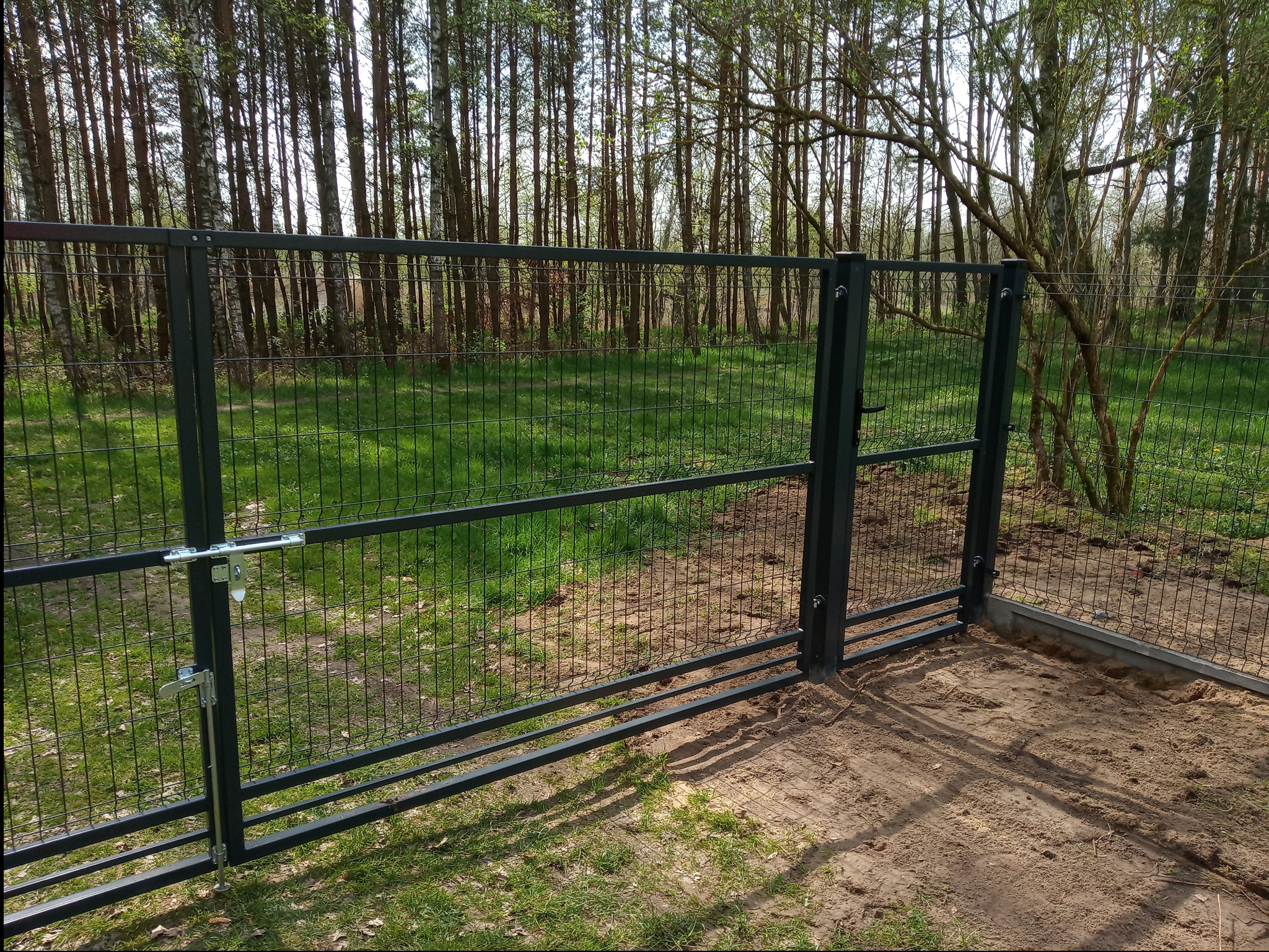 Otwarta, metalowa furtka z siatki panelowej z zamkiem na tle lasu i trawy, widoczne ślady montażu w postaci piasku i betonu.