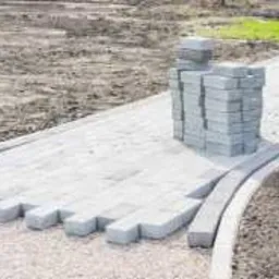 Niedokończony chodnik z szarej kostki brukowej ułożony częściowo na podsypce, obok stos kostki, widoczny krawężnik i przygotowany teren pod dalsze prace.