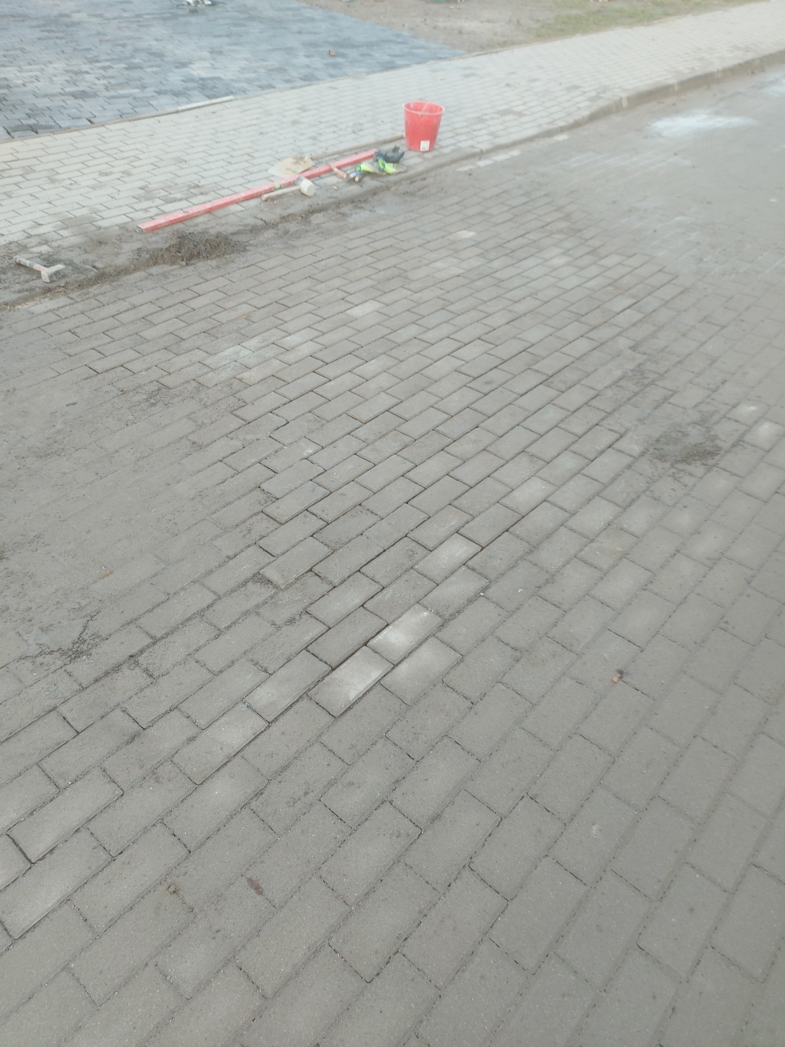 Ułożona szara kostka brukowa w trakcie prac, widoczny poziomica, młotek i czerwone wiadro na krawędzi chodnika.