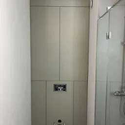 Zabudowa z płyty w odcieniach szarości, maskująca stelaż podtynkowy WC w łazience, widoczny otwór rewizyjny i przyłącze kanalizacyjne, obok kabina prysznicowa.