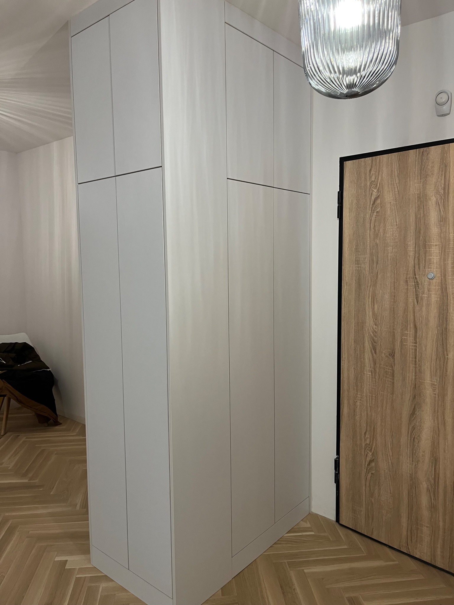 Wysoka, zabudowana szafa z szarymi, gładkimi frontami, na podłodze parkiet ułożony w jodełkę, obok drewniane drzwi wejściowe z czarną ościeżnicą i dekoracyjna lampa sufitowa.