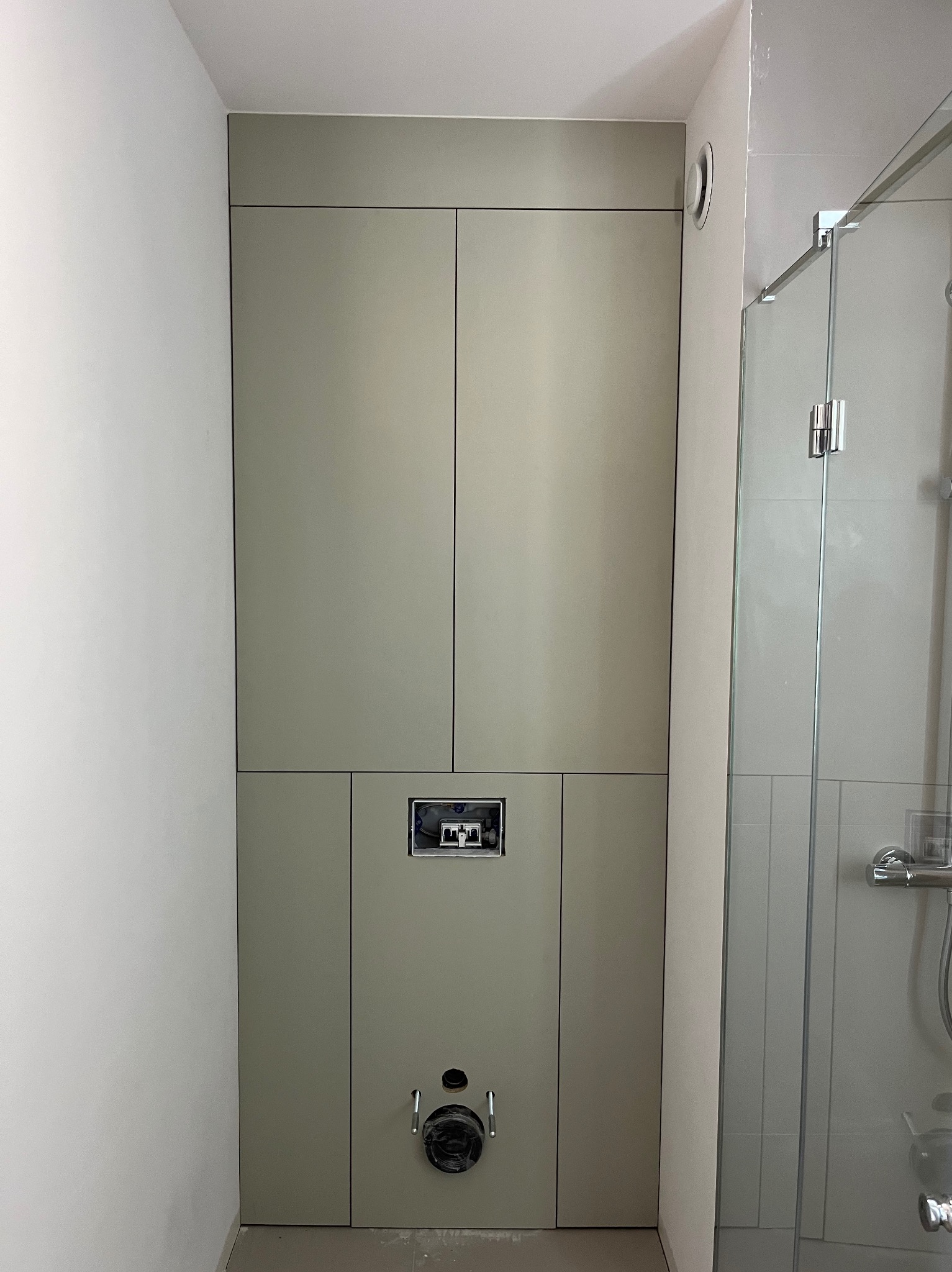 Zabudowa z płyty w odcieniach szarości, maskująca stelaż podtynkowy WC w łazience, widoczny otwór rewizyjny i przyłącze kanalizacyjne, obok kabina prysznicowa.