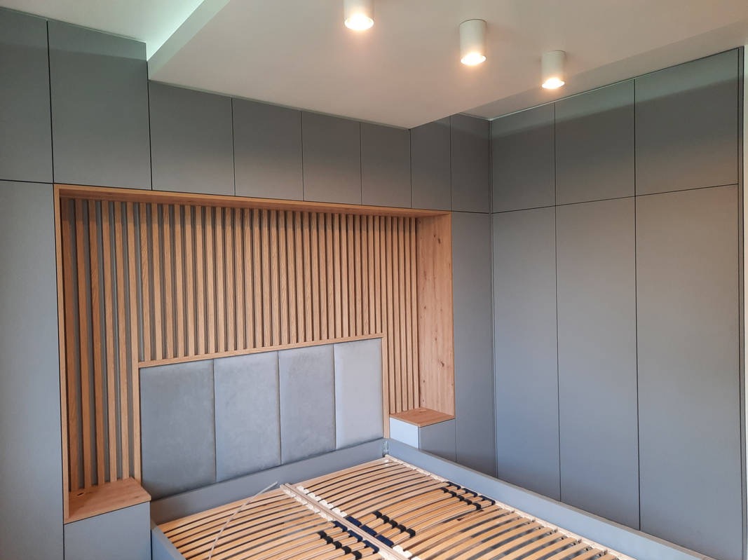 Szara szafa wnękowa z drewnianym zagłówkiem łóżka i pionowymi listwami, oświetlenie sufitowe punktowe.