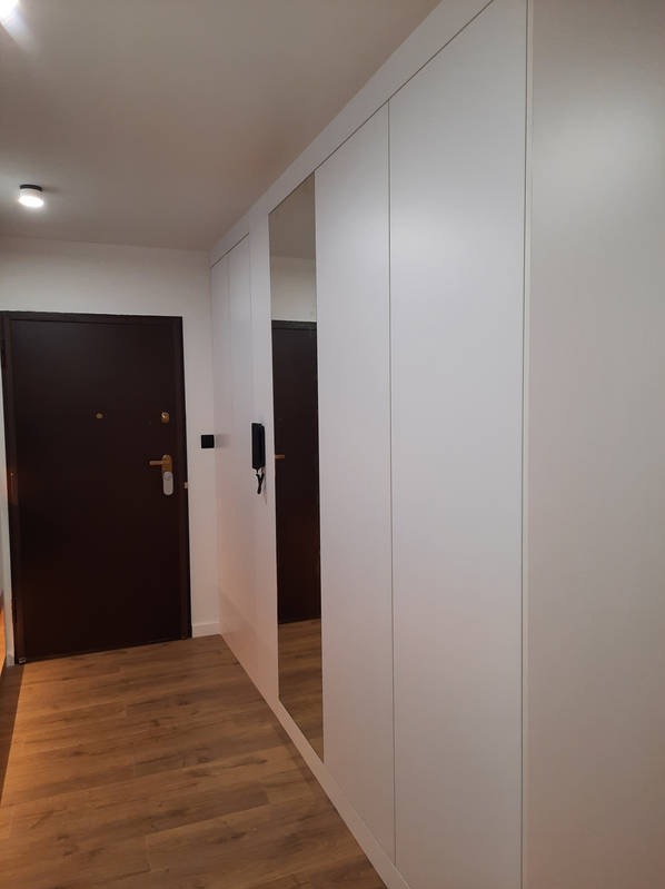 Biała szafa wnękowa z lustrem w przedpokoju, brązowe drzwi wejściowe, drewnopodobna podłoga, minimalistyczny design.