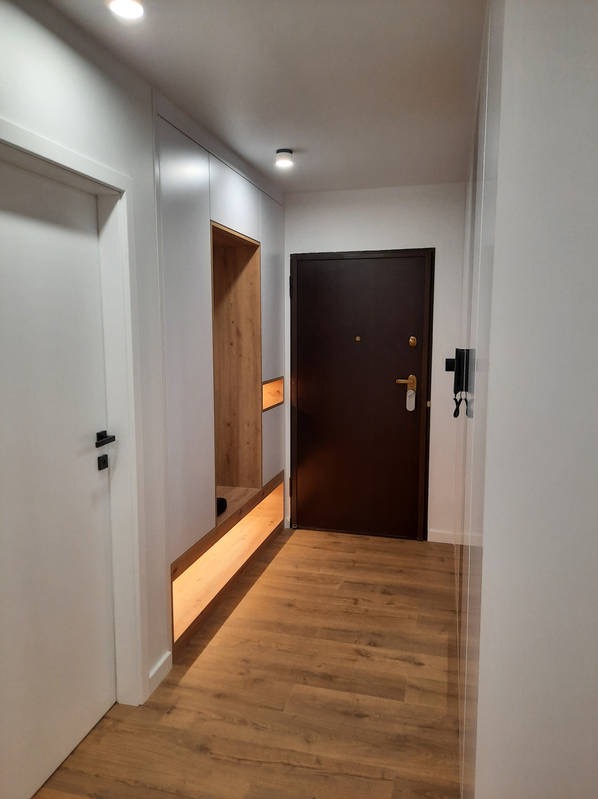 Nowoczesny przedpokój z białą szafą wnękową z elementami drewnopodobnymi i podświetleniem LED, brązowe drzwi wejściowe, podłoga z paneli imitujących drewno.