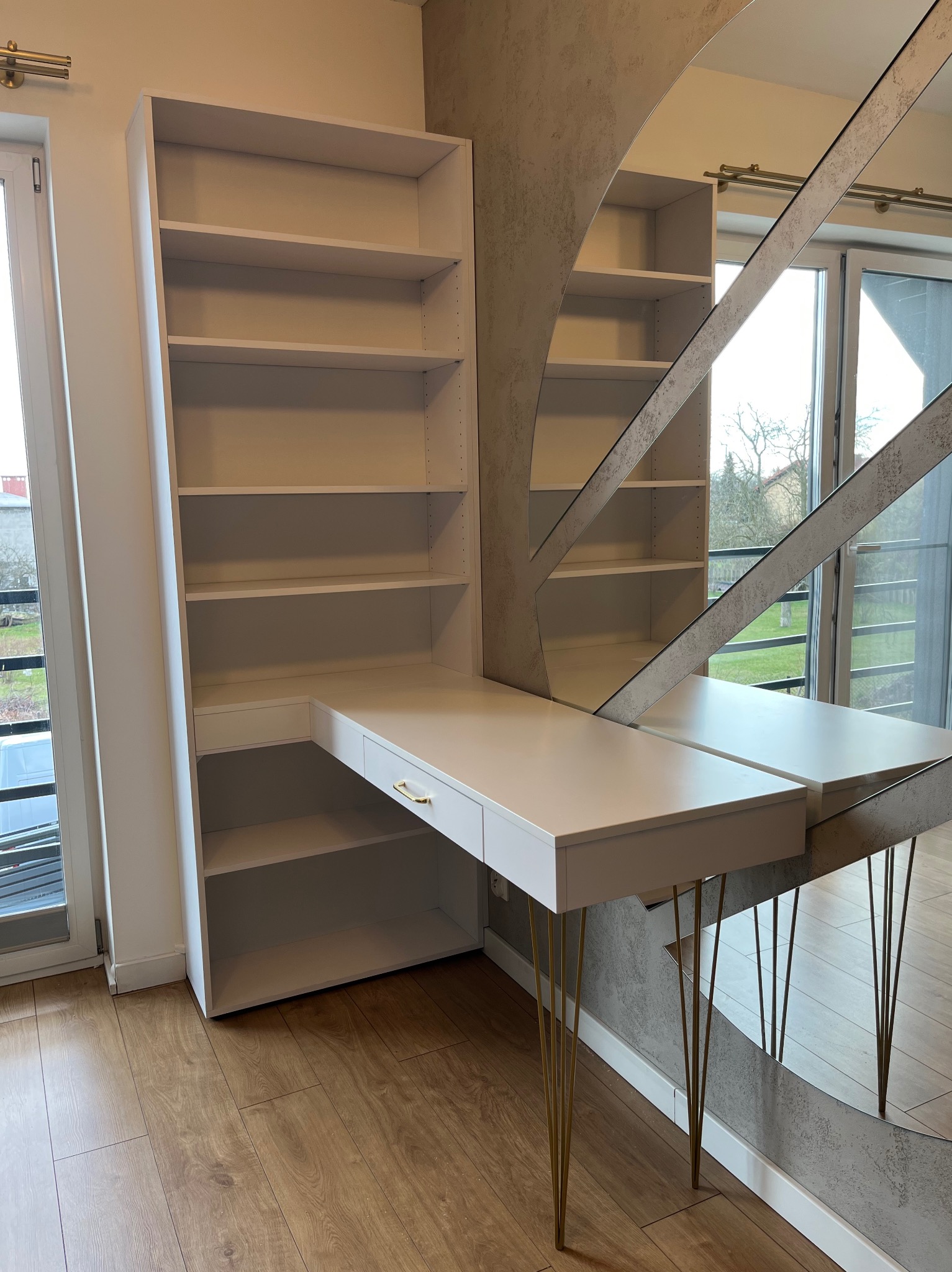 Białe biurko z regałem połączone z toaletką z owalnym lustrem w metalowej ramie, wsparte na złotych nóżkach w jasnym pokoju.