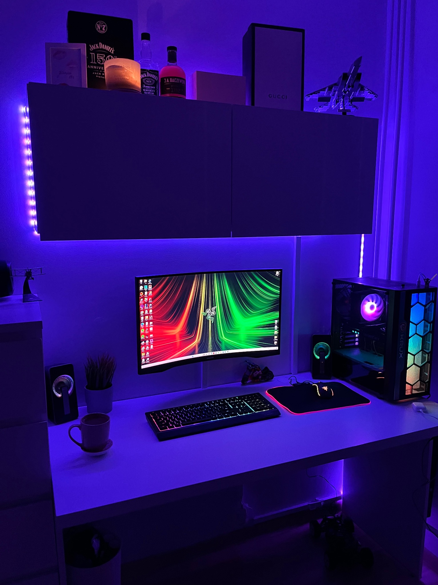 Pokój gracza z biurkiem podwieszanym, oświetleniem LED, komputerem stacjonarnym z podświetlaną obudową, monitorem z tapetą Razer, klawiaturą, myszką, głośnikami i dekoracjami na szafce.