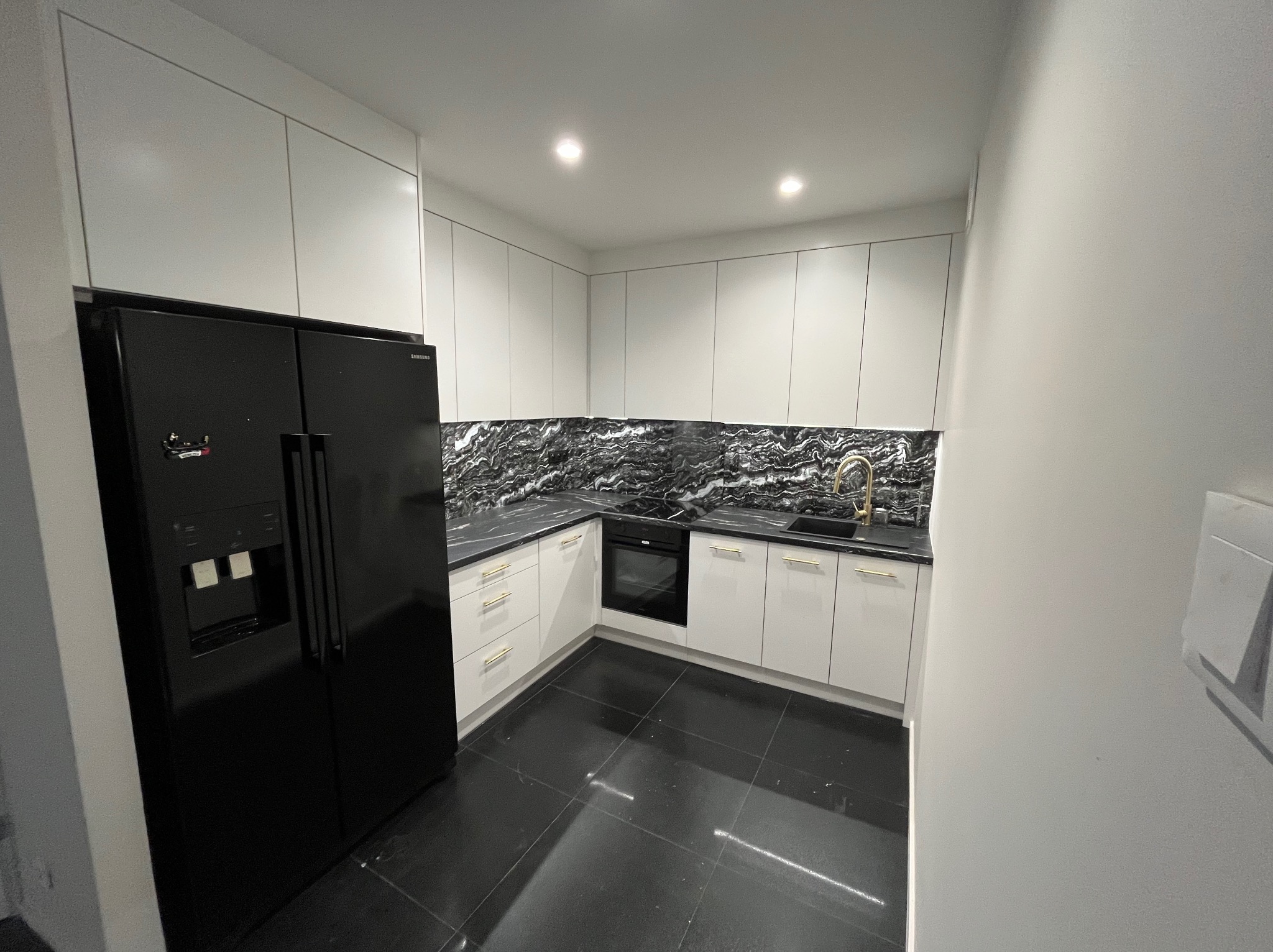 Nowoczesna kuchnia z białymi szafkami, czarnym blatem imitującym marmur, złotą armaturą i czarną lodówką side-by-side.