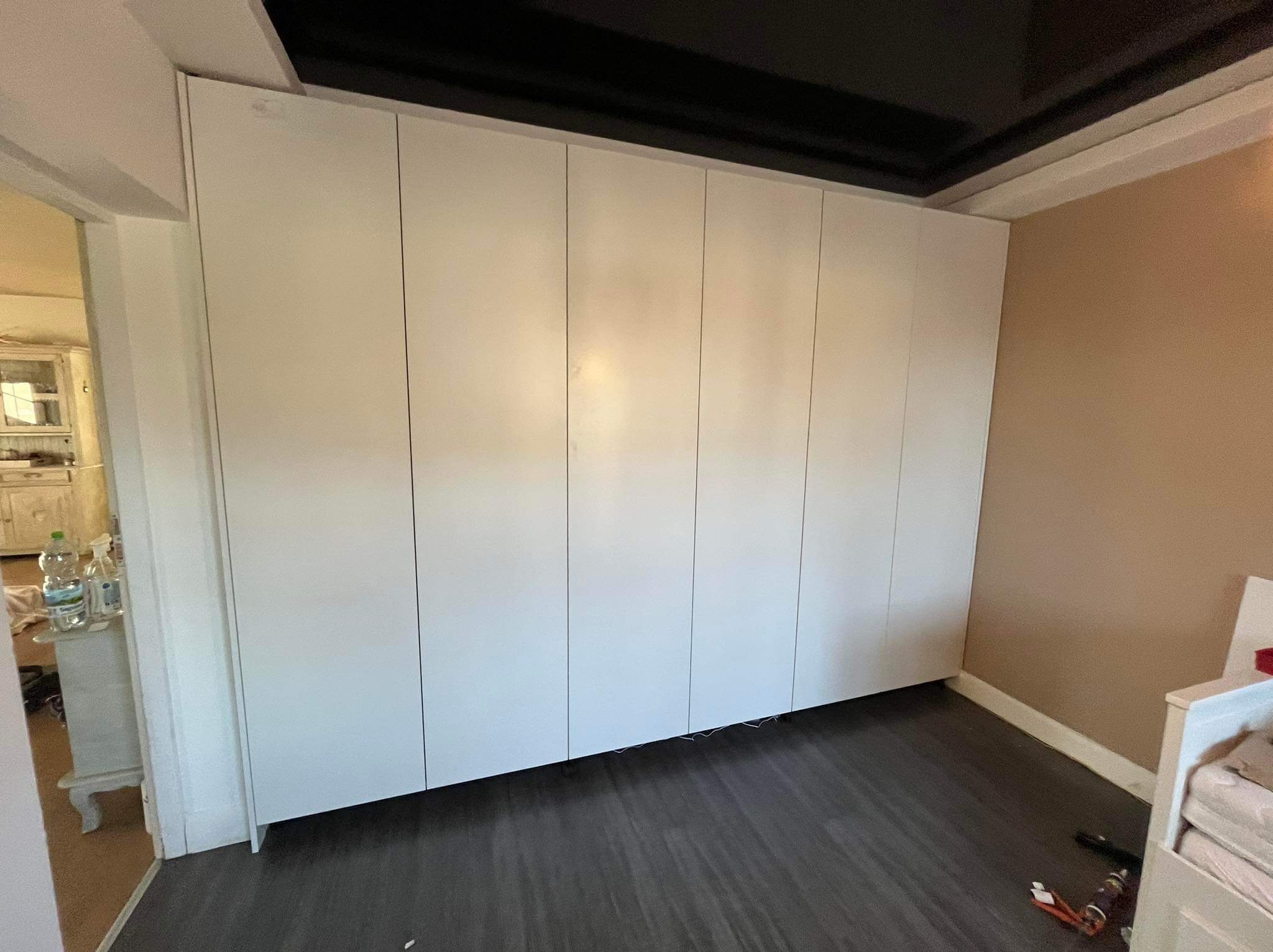 Biała szafa wnękowa z siedmioma drzwiami, widok z perspektywy podłogi, fragment pokoju z ciemną podłogą i beżową ścianą, widoczny fragment pokoju obok z meblami i butelkami.
