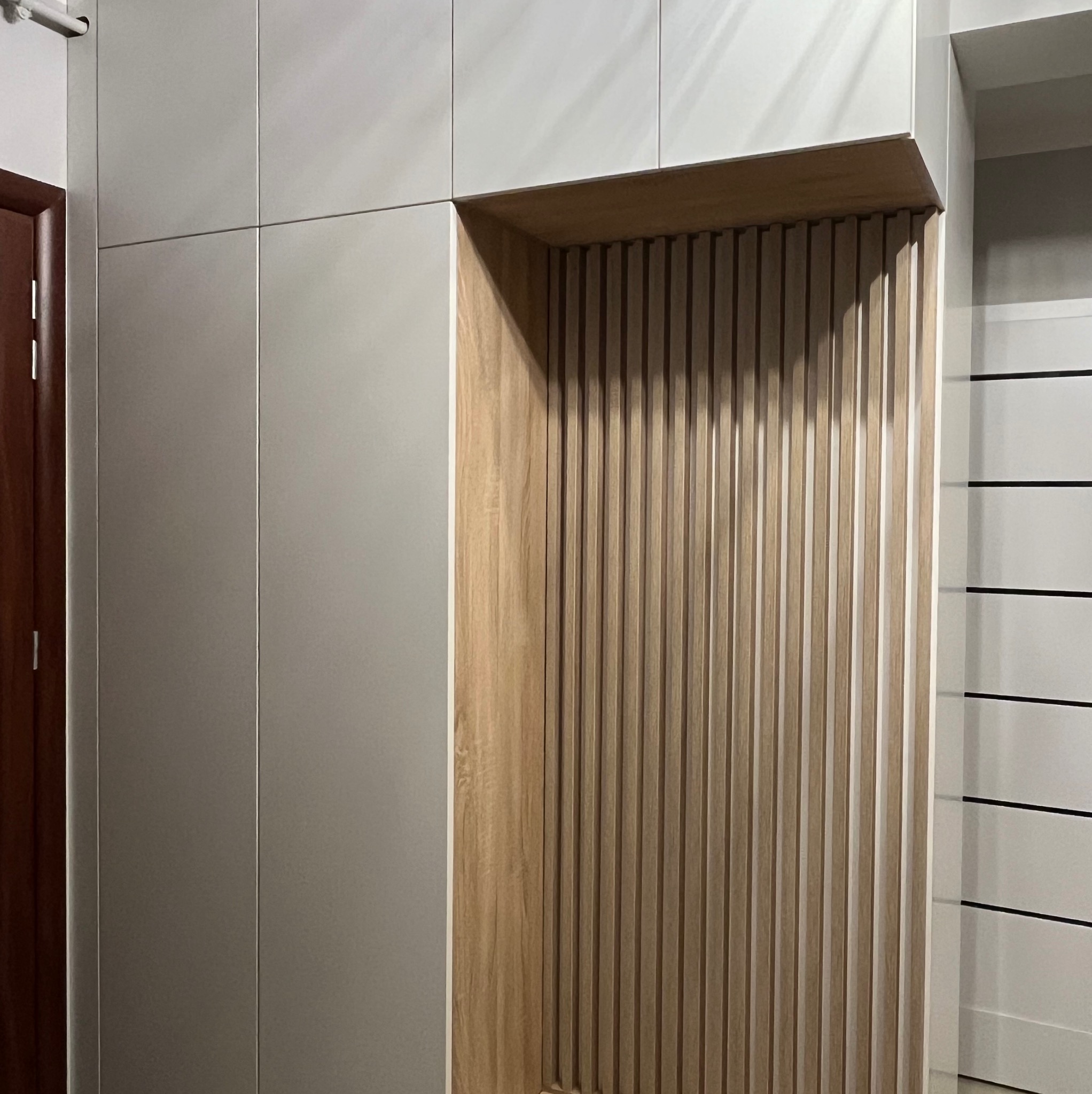 Szara szafa wnękowa z drewnianą wnęką z pionowymi listwami, widoczny fragment drzwi wejściowych i element białej ściany z czarnymi poziomymi paskami.