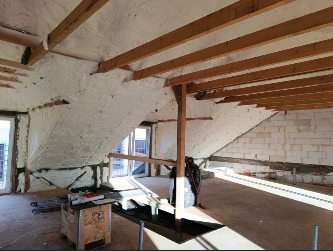 Poddasze z drewnianą konstrukcją dachu i ścianami pokrytymi białą pianą izolacyjną, widoczne fragmenty muru z białych bloczków oraz okna, na podłodze rozłożone materiały budowlane i skrzynia...