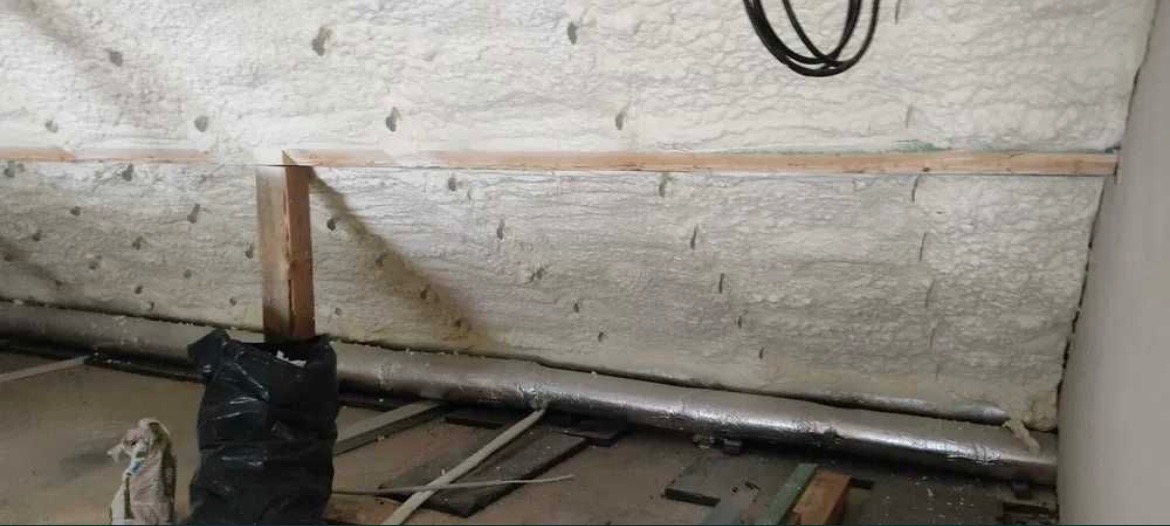 Ściana pokryta szarą pianą izolacyjną z widocznymi śladami po wtryskiwaniu, drewniana listwa mocująca, srebrna rura w izolacji i czarny worek na podłodze.