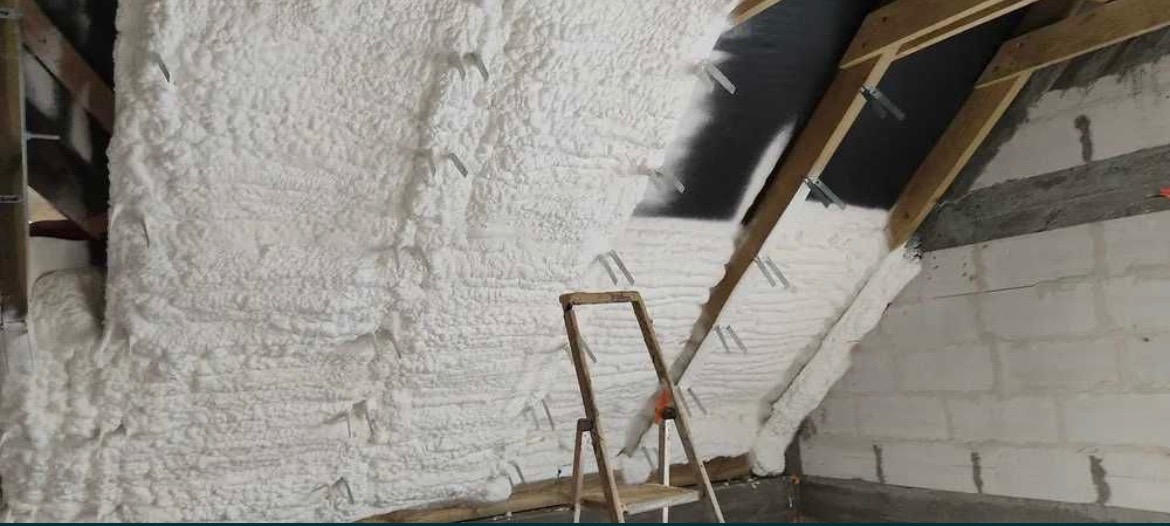 Poddasze w trakcie izolacji: biała pianka pokrywa ściany i krokwie, widoczne zszywki mocujące, drewniana drabina oparta o ścianę z pustaków.