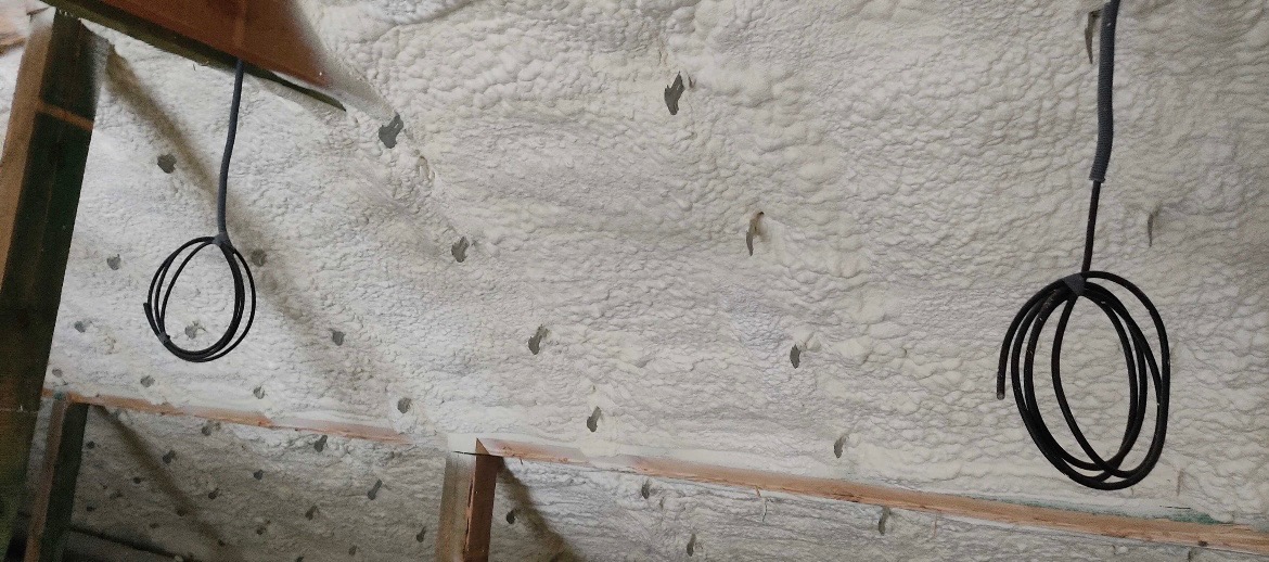 Świeżo nałożona warstwa izolacji piankowej z widocznymi przewodami elektrycznymi, przymocowanymi do sufitu. Widoczne drewniane elementy konstrukcyjne dachu.