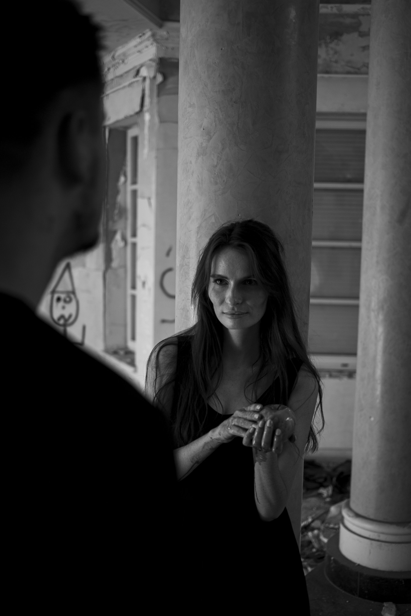 Czarno-białe zdjęcie kobiety opierającej się o kolumnę w opuszczonym budynku, trzymającej w dłoniach jabłko, z rozmazaną postacią mężczyzny w lewym dolnym rogu kadru.