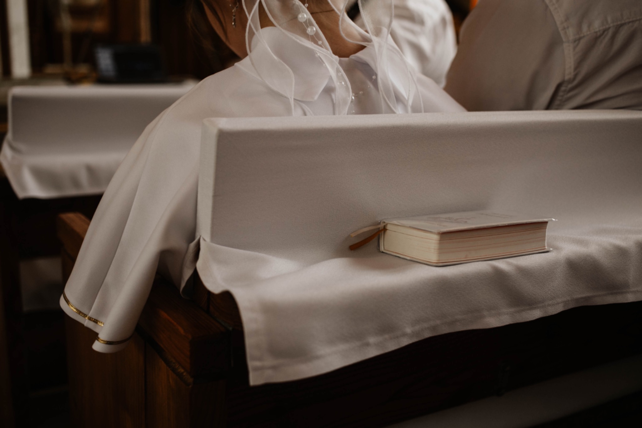 Dziewczynka w białej albie i welonie klęczy w ławce kościelnej, obok leży modlitewnik z zakładką.