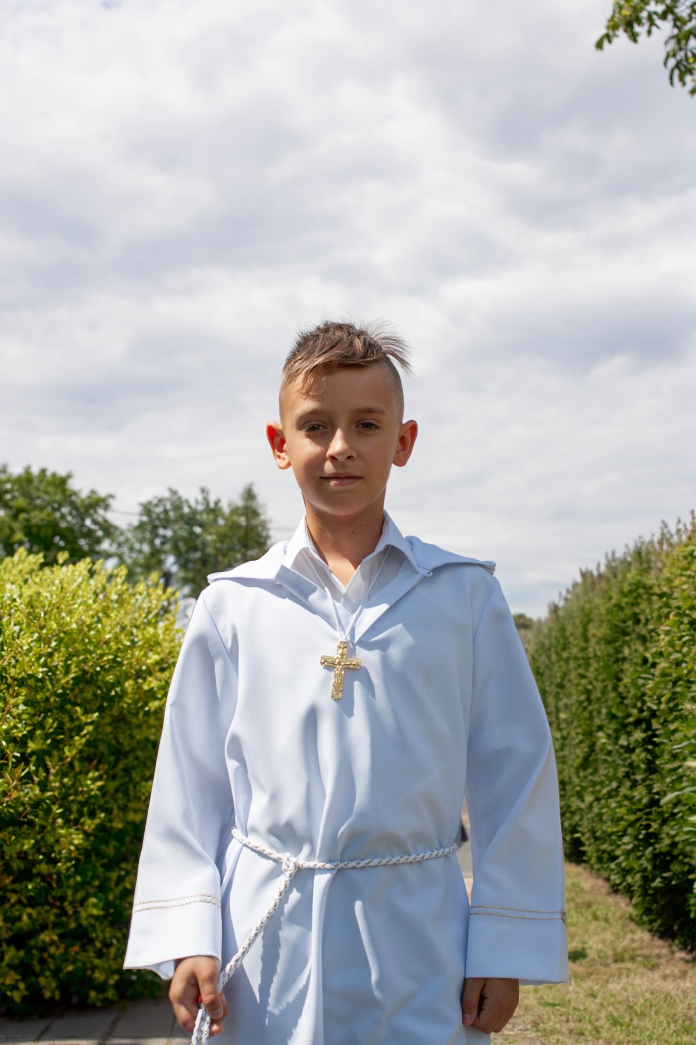 Chłopiec w stroju komunijnym z złotym krzyżykiem na tle zieleni i pochmurnego nieba.