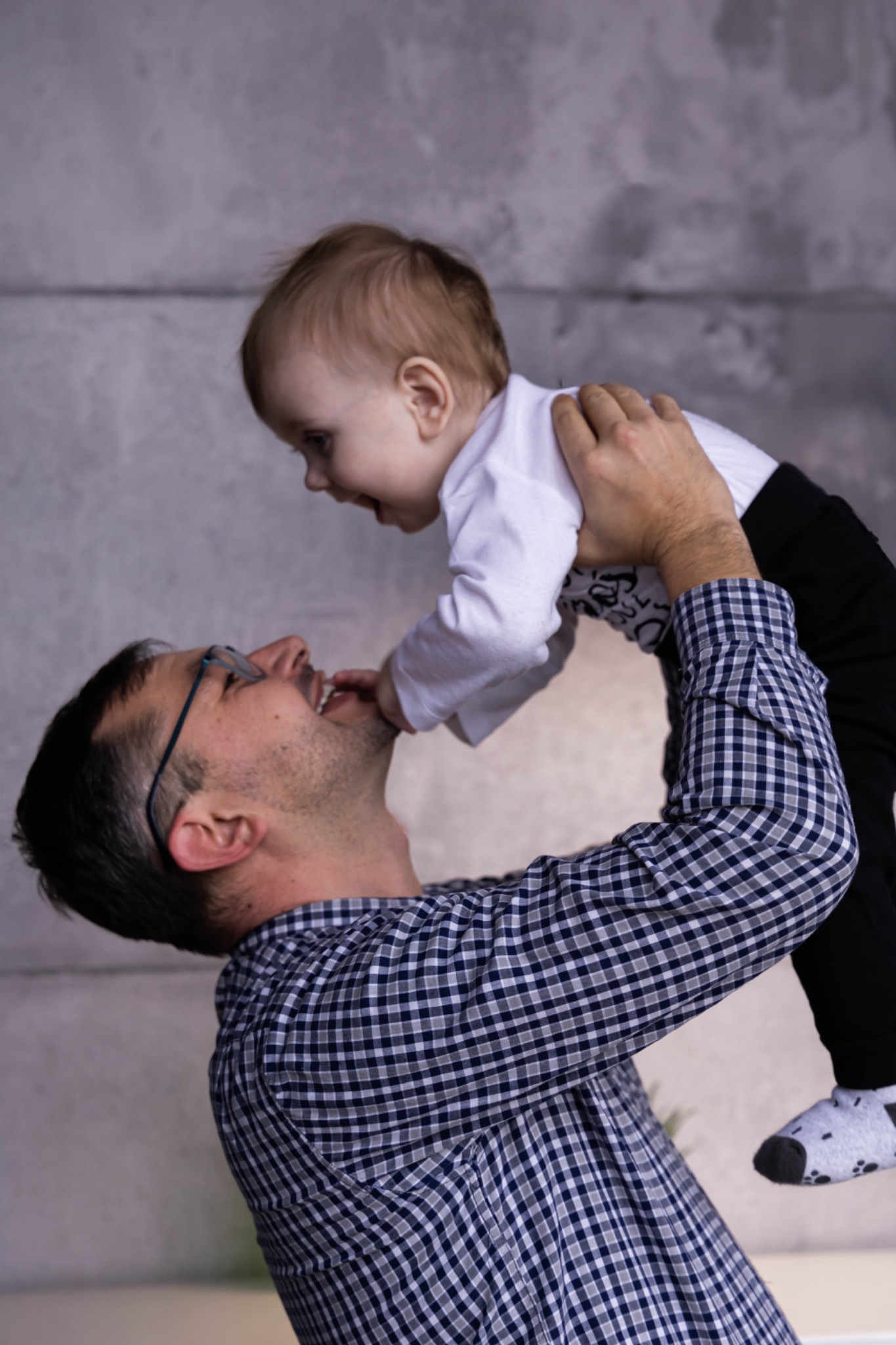 Uśmiechnięty tata w okularach podnosi do góry roześmiane dziecko na tle szarej ściany.