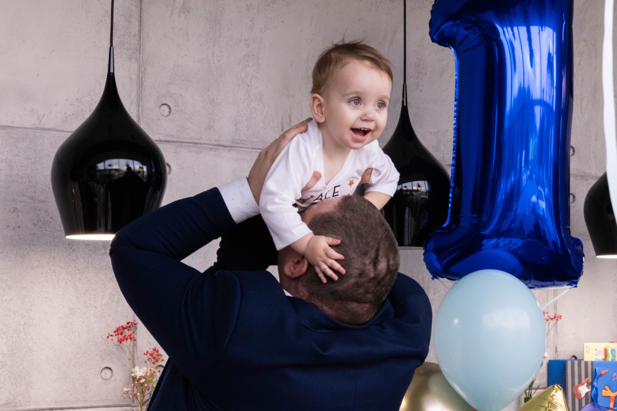 Uśmiechnięty roczny chłopiec w białym ubranku uniesiony przez ojca w niebieskim garniturze na tle betonowej ściany z czarnymi lampami i niebieskim balonem w kształcie cyfry 1.