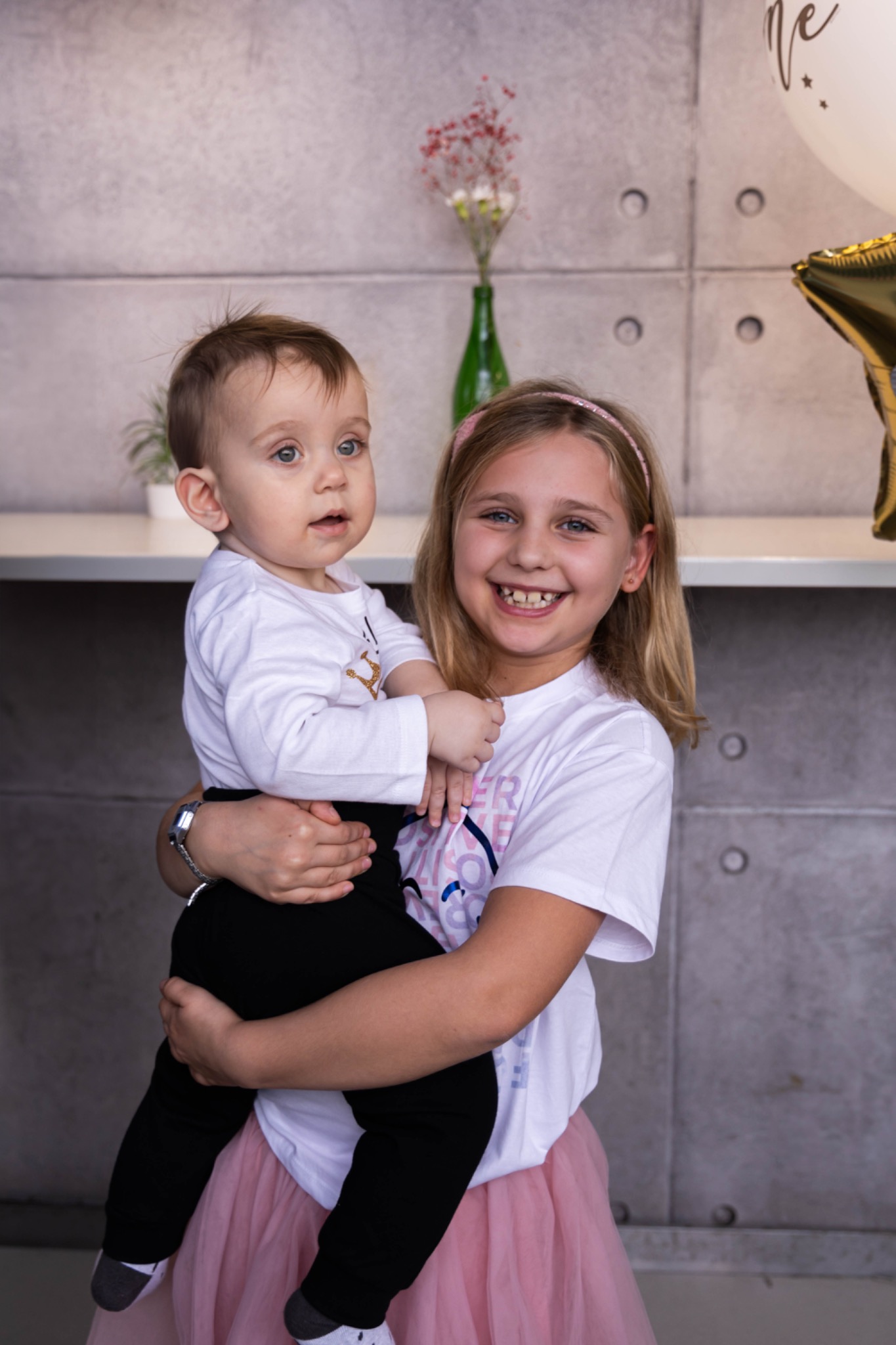 Uśmiechnięta dziewczynka w różowej tiulowej spódnicy trzyma na rękach młodszego brata w białej koszulce i czarnych spodniach na tle betonowej ściany z dekoracjami.