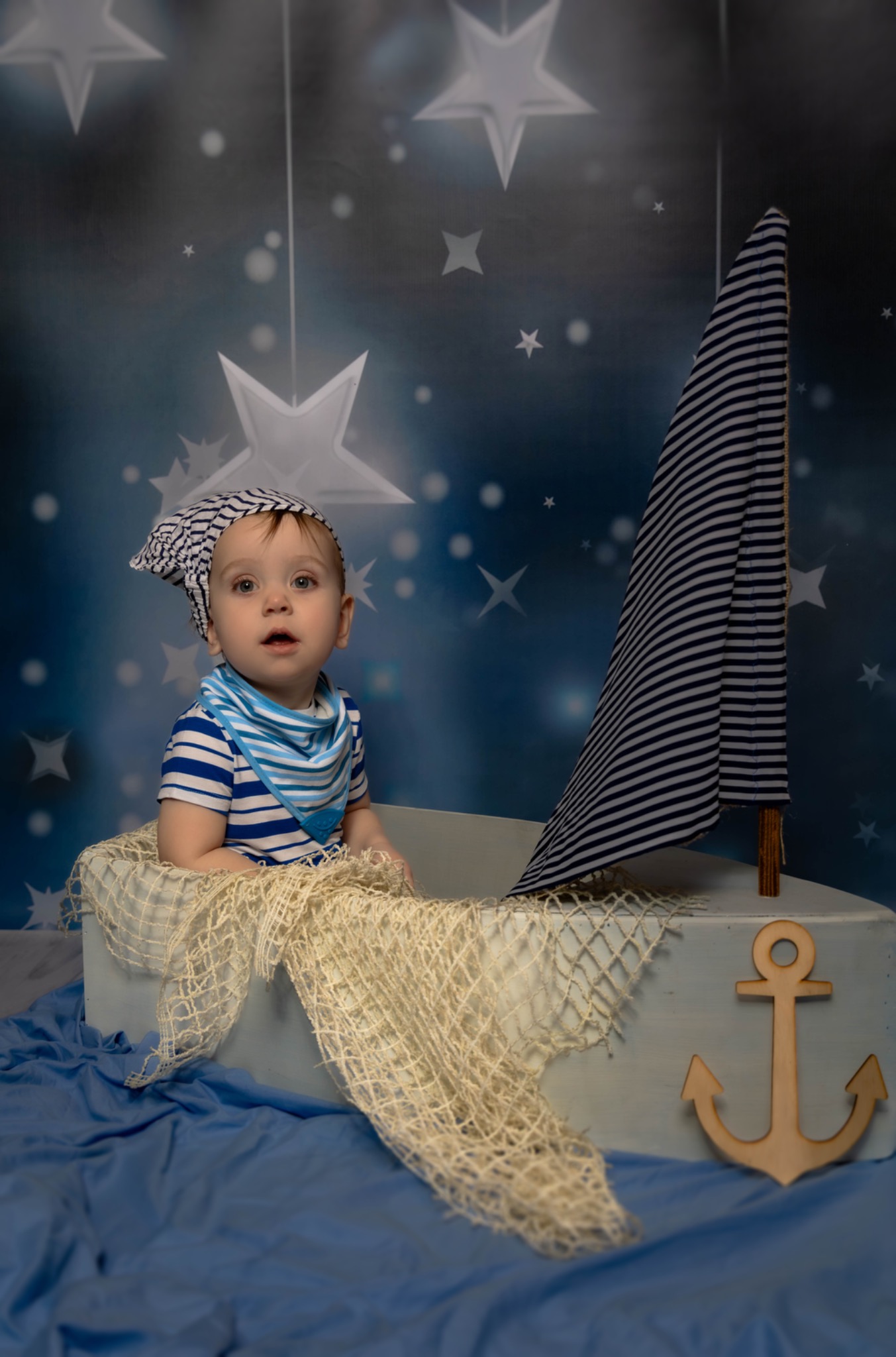 Uroczy niemowlak w marynarskim stroju i czapce siedzi w dekoracyjnej łódce z żaglem na tle gwieździstego nieba, stylizowany na małego żeglarza.