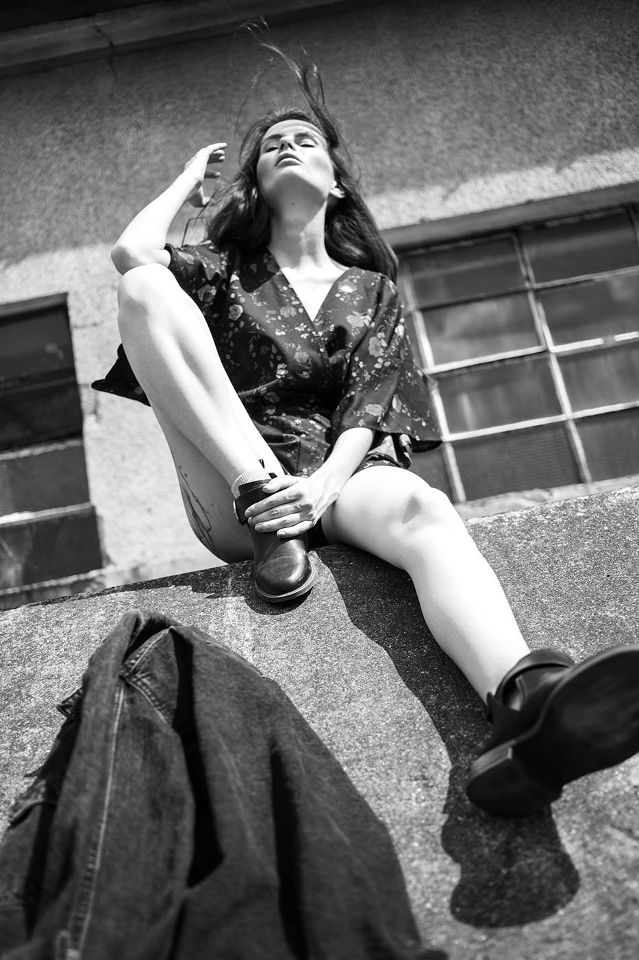 Czarno-białe zdjęcie kobiety siedzącej na betonowym murku w kwiecistej bluzce i butach, z rozwianymi włosami i zamkniętymi oczami, z denimową kurtką obok, sfotografowane z dołu na tle budynku...