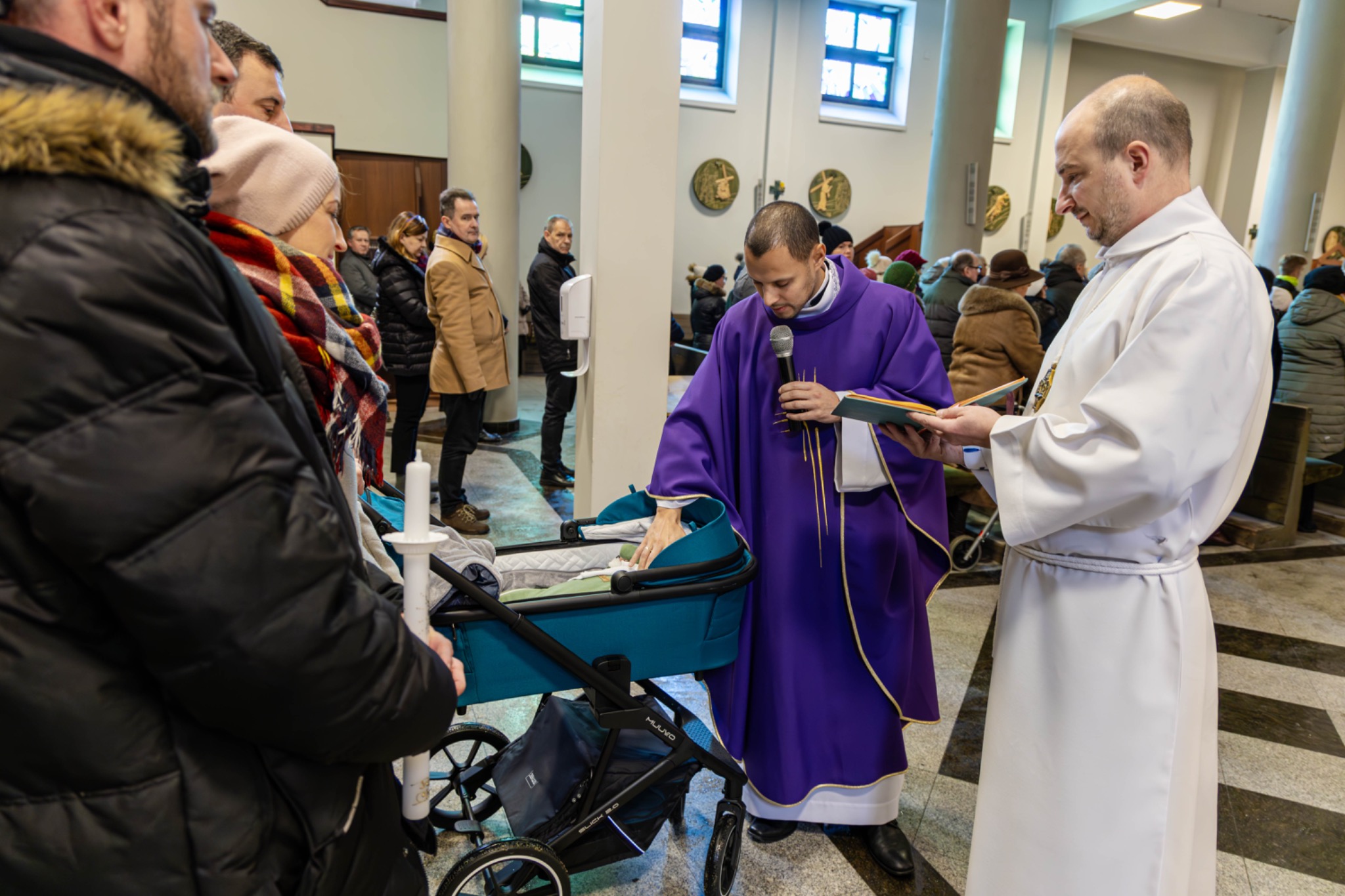 Ksiądz w fioletowej szacie z mikrofonem błogosławi dziecko w turkusowym wózku podczas ceremonii w kościele, obok stoi drugi ksiądz w białej szacie z księgą, w tle zgromadzeni ludzie.