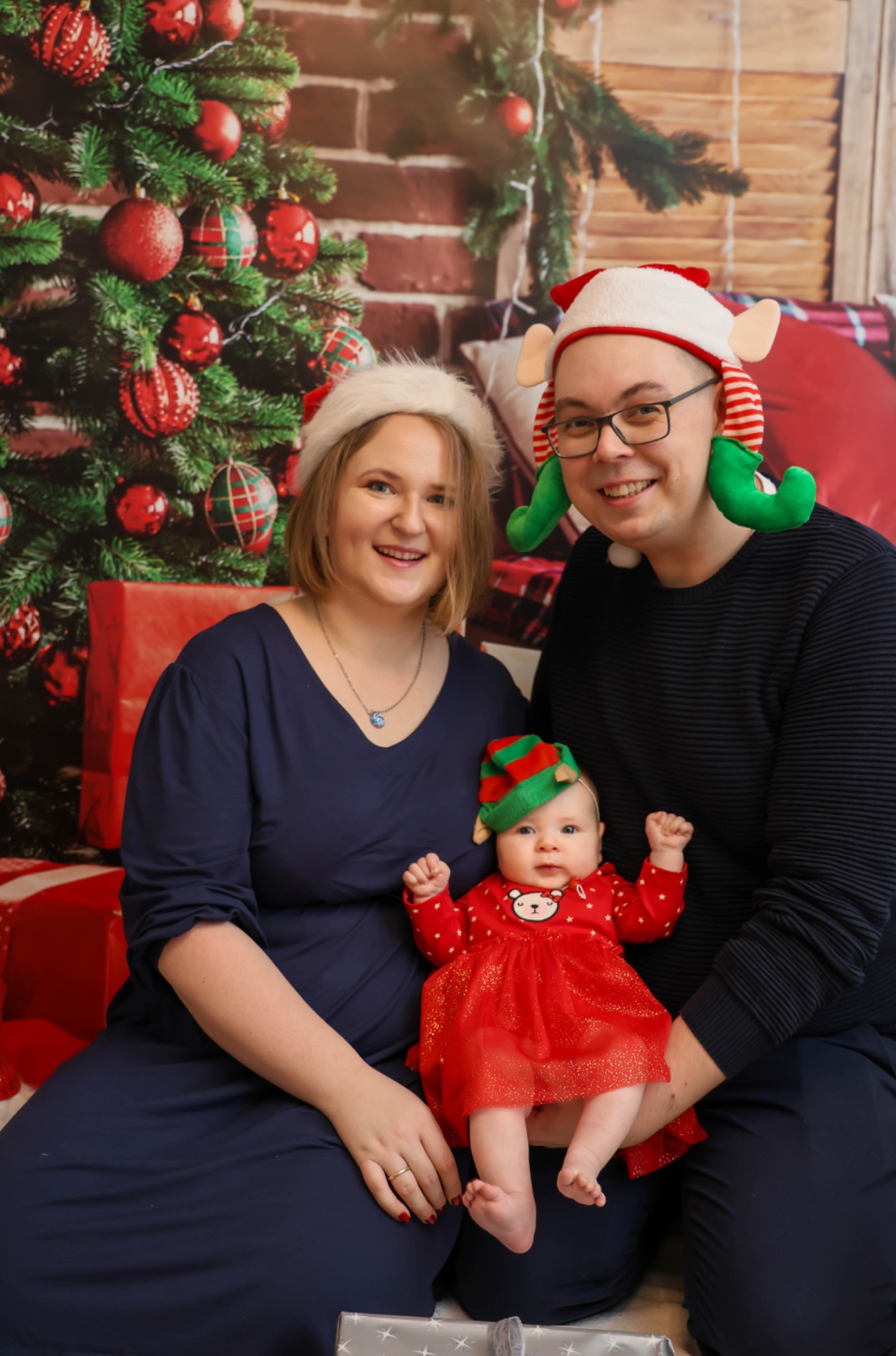Rodzinne zdjęcie w świątecznej scenerii: rodzice w czapkach Mikołaja i elfa trzymają ubraną w czerwoną sukienkę z motywem misia córeczkę na tle bożonarodzeniowej dekoracji z prezentami.