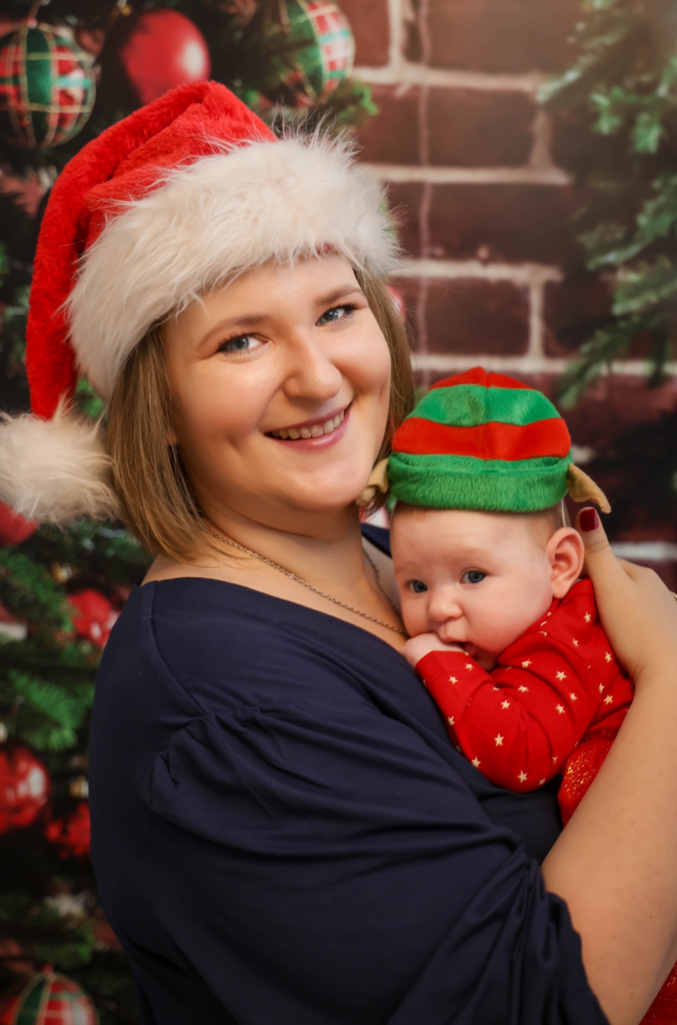Uśmiechnięta kobieta w czapce Świętego Mikołaja trzyma na rękach niemowlę w czerwonym ubranku i czapce elfa na tle dekoracji świątecznych.