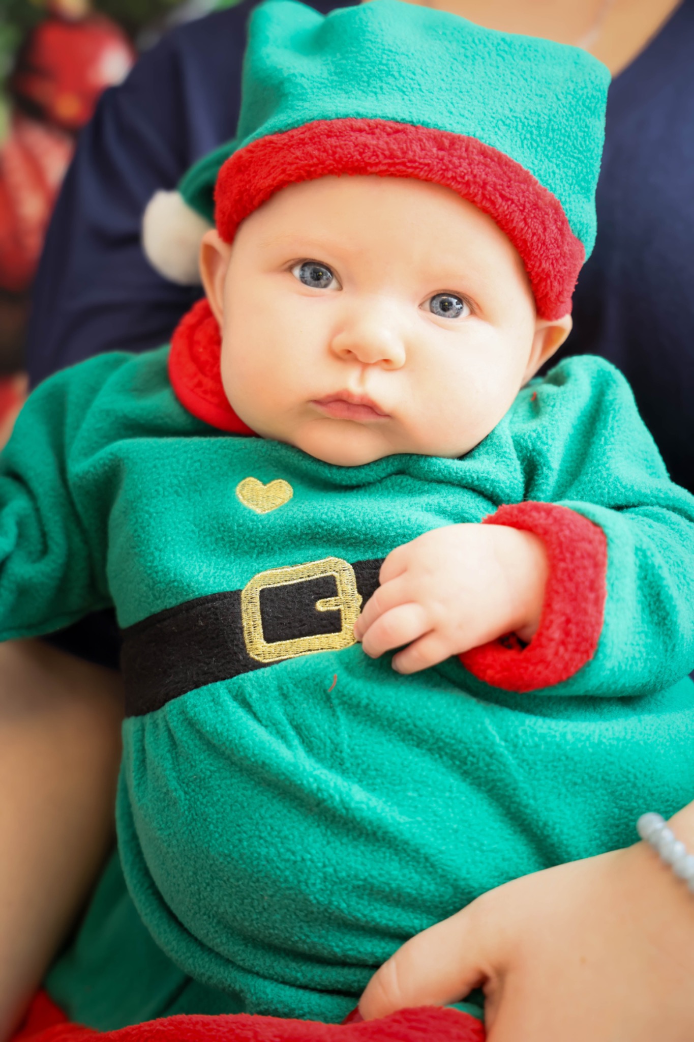 Niemowlę z jasnymi oczami ubrane w zielony strój elfa z czerwonymi akcentami i czarnym pasem z złotą klamrą, siedzące na kolanach osoby dorosłej.