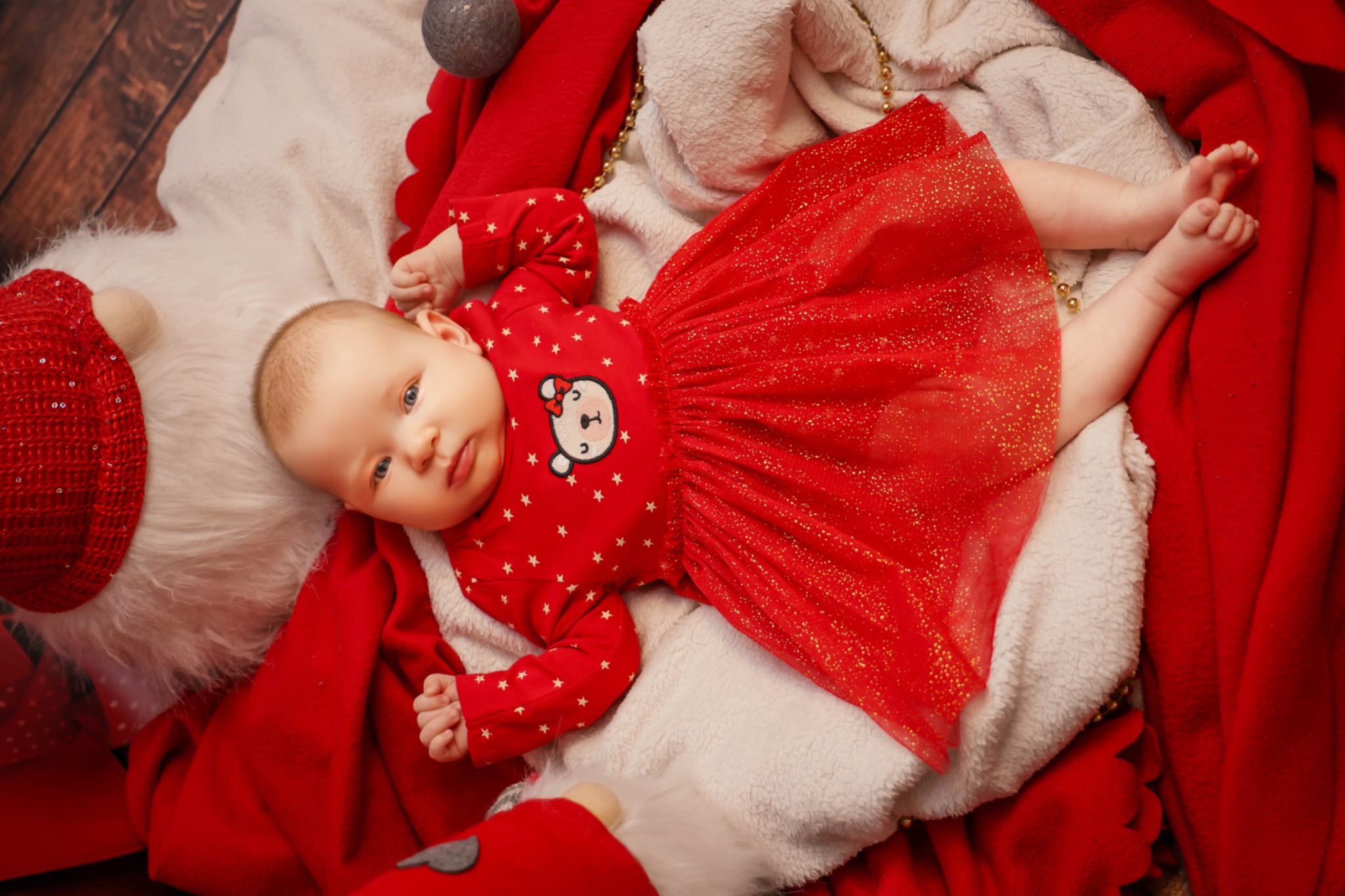 Niemowlę w czerwonej sukience ze wzorem gwiazdek i misia, leżące na białym futerku i czerwonym materiale, z elementami dekoracji świątecznych.