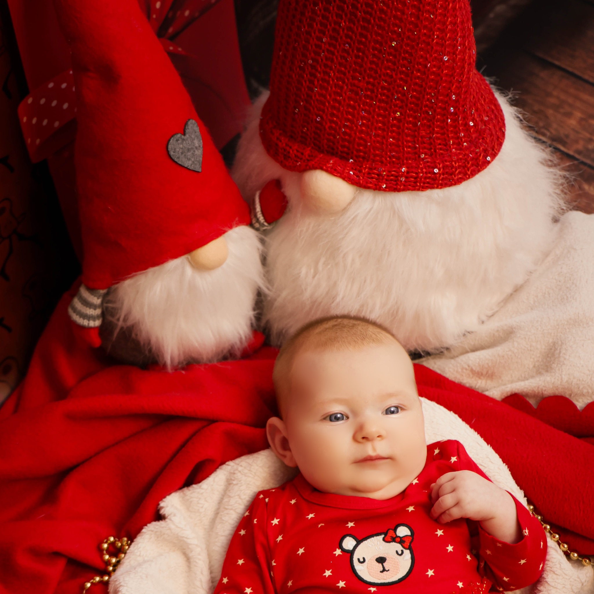 Niemowlę w czerwonym ubranku z motywem misia leży na białym kocyku, otoczone czerwonym materiałem i dekoracyjnymi gnomami w czerwonych czapkach.