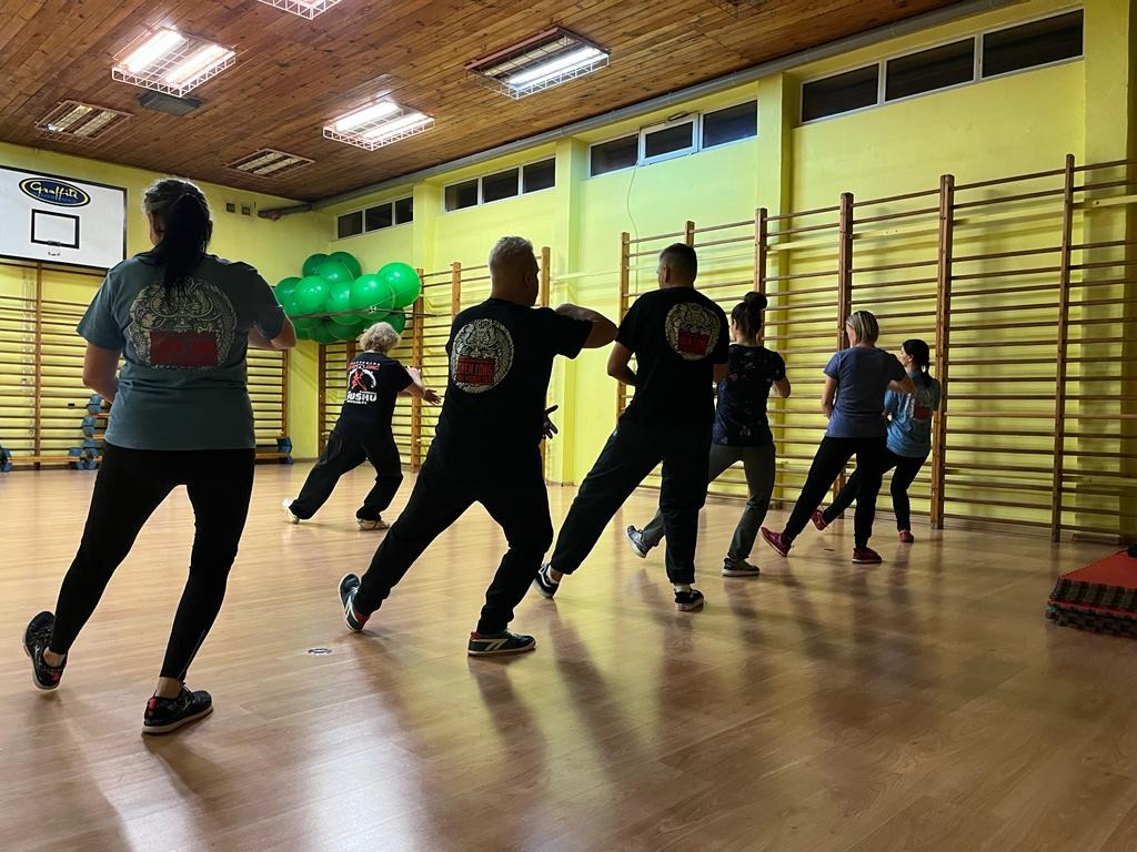 Grupa osób ćwiczy w sali gimnastycznej, wykonując sekwencję ruchów charakterystycznych dla sportów walki; widoczne drabinki gimnastyczne na ścianie i dekoracja z zielonych balonów.