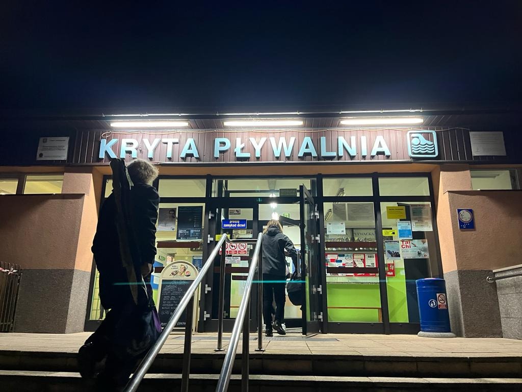 Wejście do krytej pływalni nocą, dwie osoby wchodzą po schodach, jedna z futerałem na instrument muzyczny na plecach, widoczne plakaty i informacje na drzwiach.