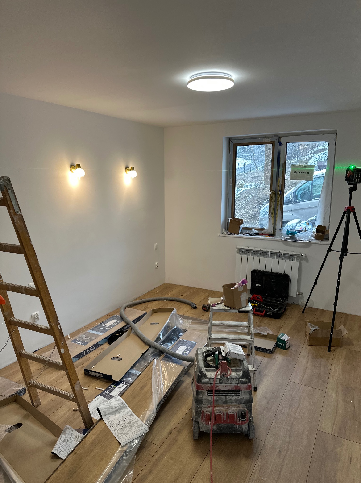 Pokój w trakcie układania paneli podłogowych. Widoczne rozłożone panele, drabina, odkurzacz przemysłowy i poziomica laserowa na statywie.