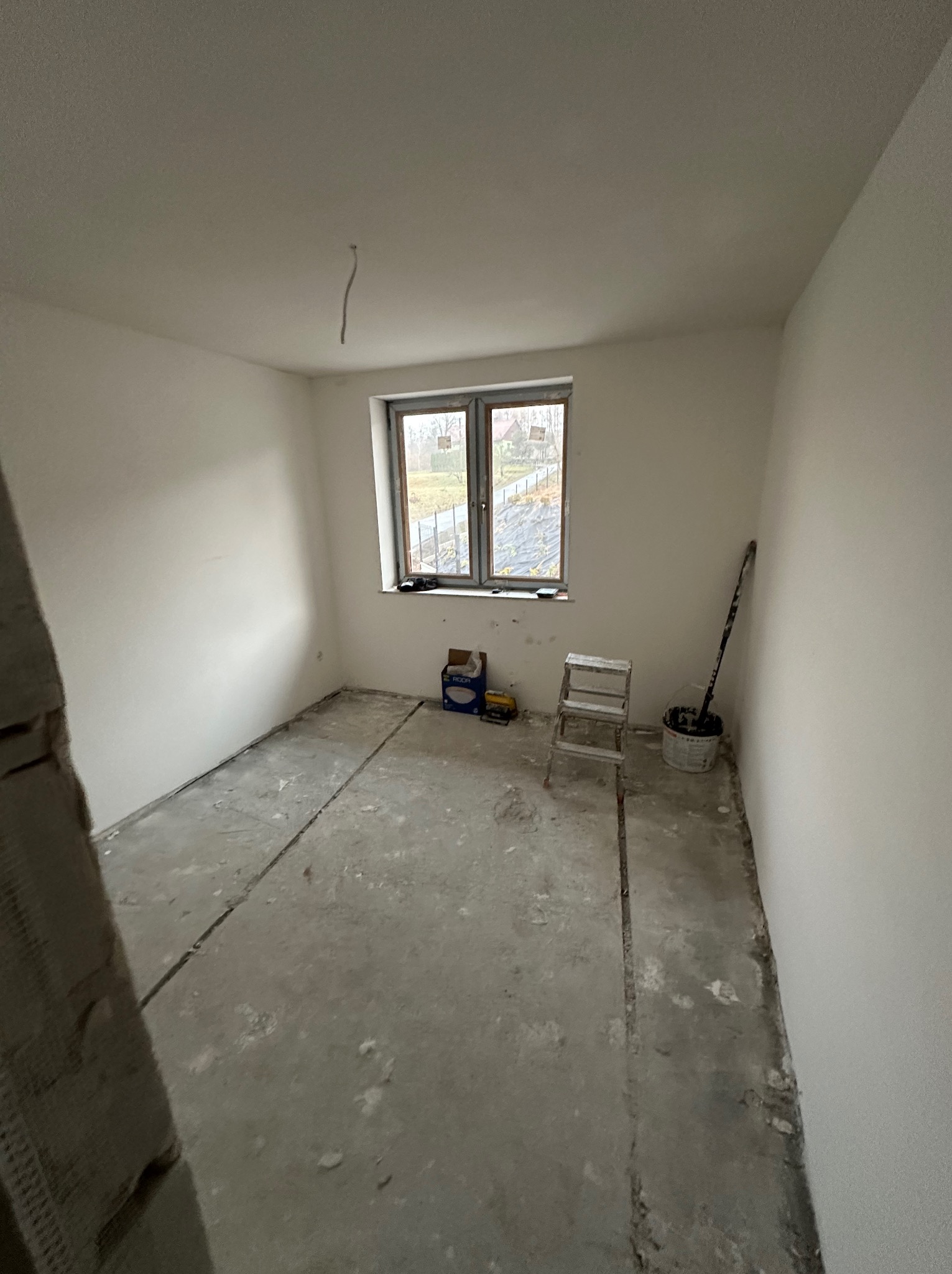 Pokój w trakcie remontu: świeżo wyszpachlowane ściany, surowa podłoga, drabina malarska, wiadro z farbą i okno z widokiem na zewnątrz. Widoczne ślady prac wykończeniowych.