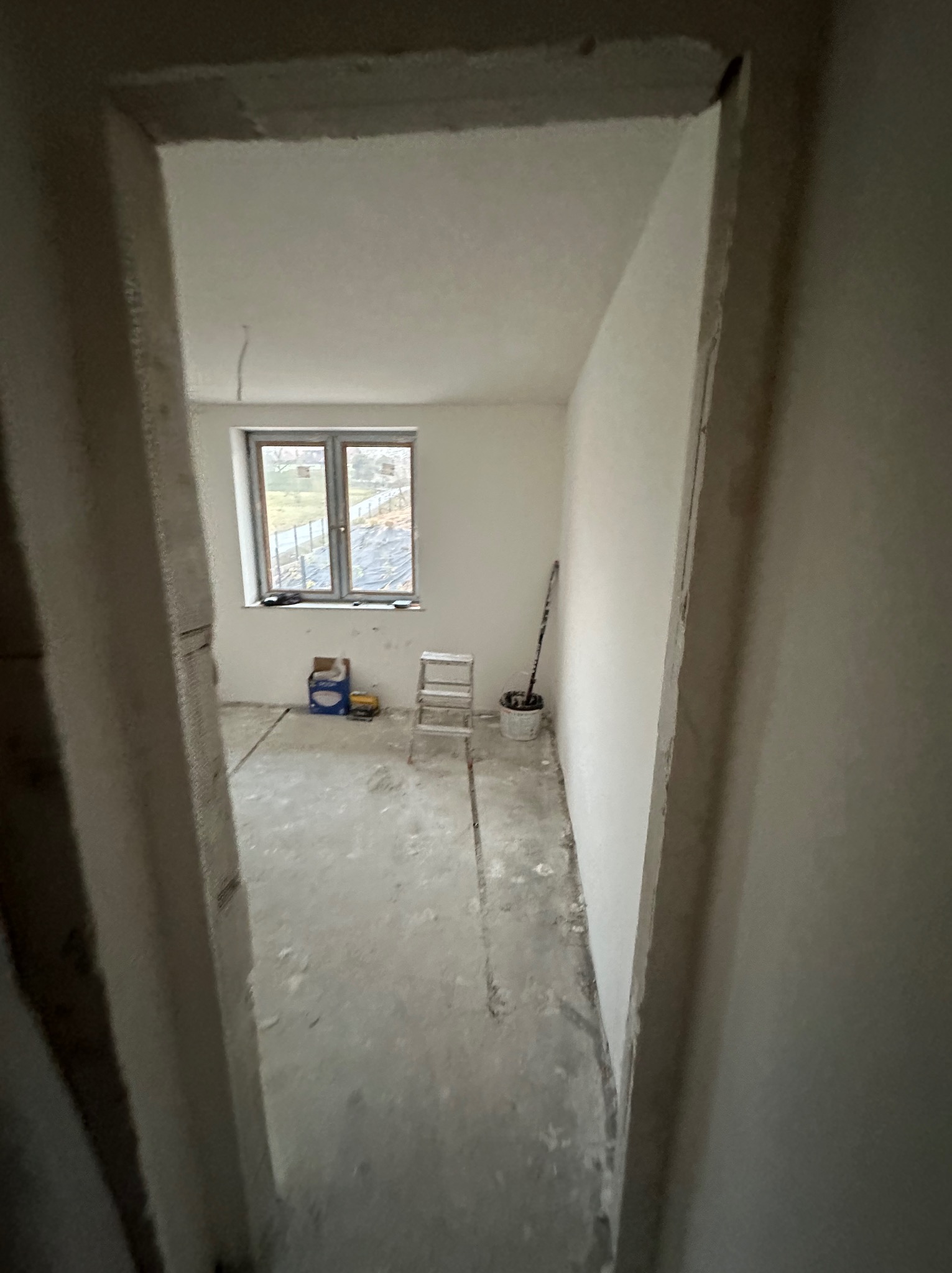 Surowy stan wnętrza w trakcie remontu, widok przez futrynę. Ściany przygotowane do gładzi, podłoga surowa, okno z widokiem na zewnątrz. W tle drabina i narzędzia.