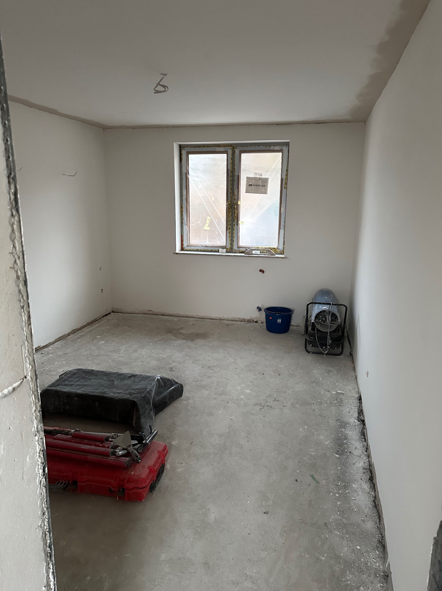 Pokój w trakcie remontu: świeżo otynkowane ściany, okno z naklejkami, narzędzia w czerwonej skrzynce, nagrzewnica i niebieskie wiadro. Widok zza framugi.