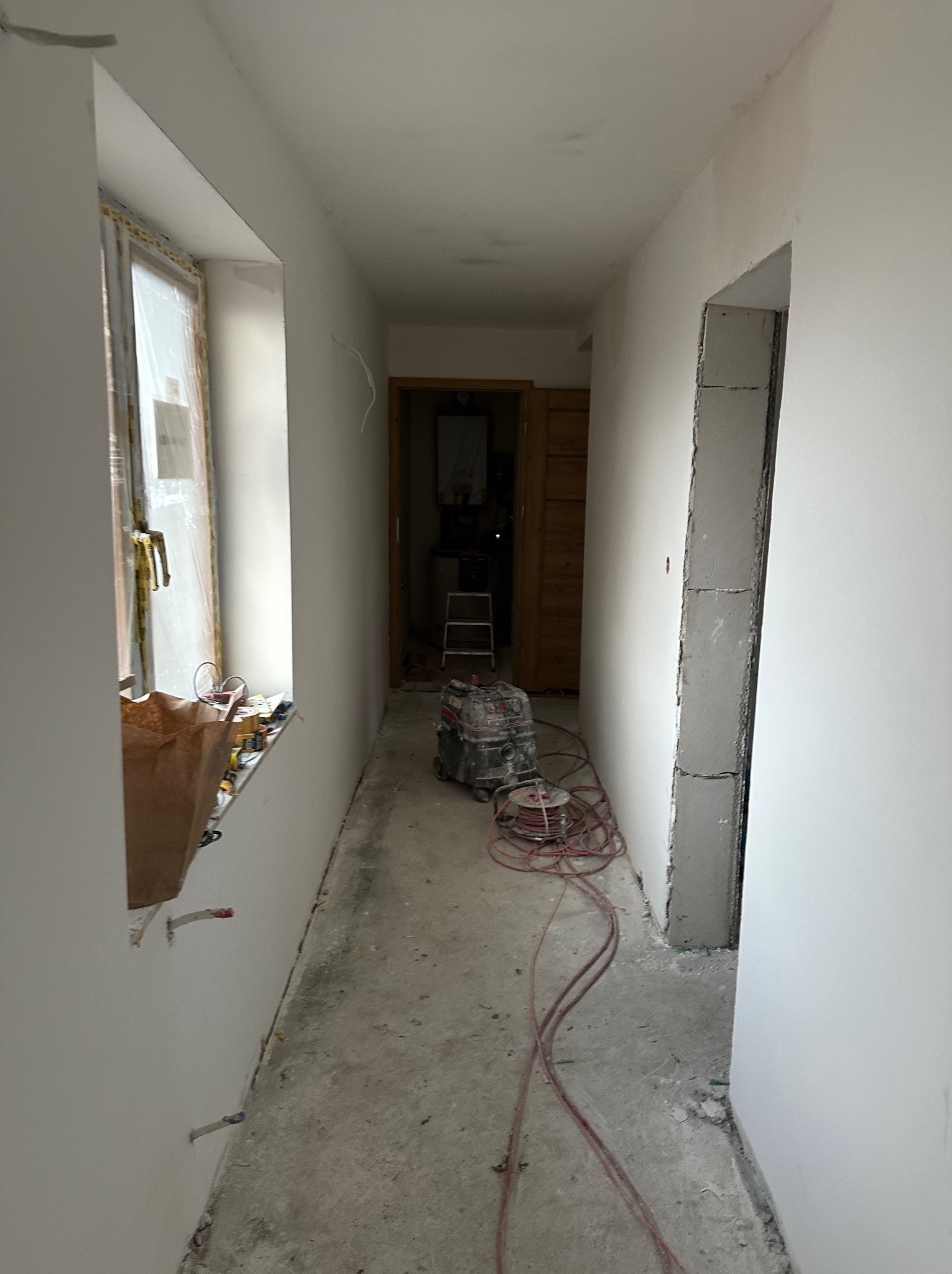 Korytarz w trakcie remontu: świeżo pomalowane ściany na biało, widoczne narzędzia i materiały budowlane, surowy beton przy otworze drzwiowym, okno z folią ochronną.
