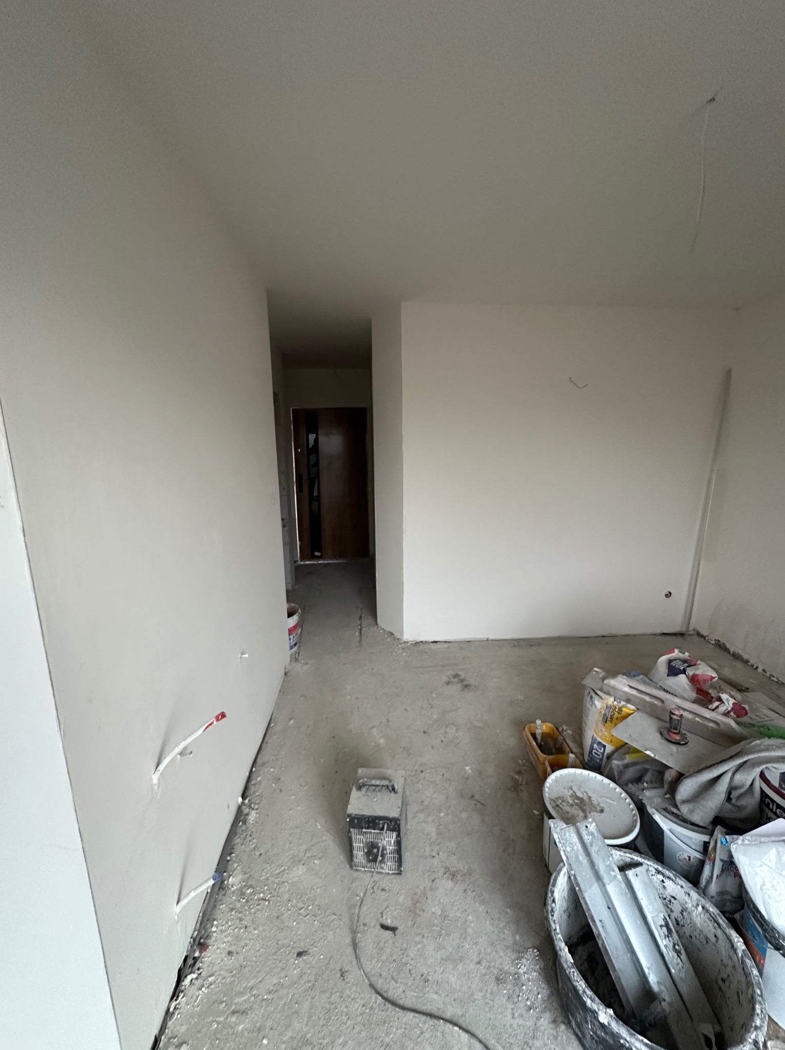 Wnętrze po szpachlowaniu ścian, widoczne narzędzia i materiały budowlane. Ściany przygotowane do dalszych prac wykończeniowych. Widok na korytarz z drzwiami.
