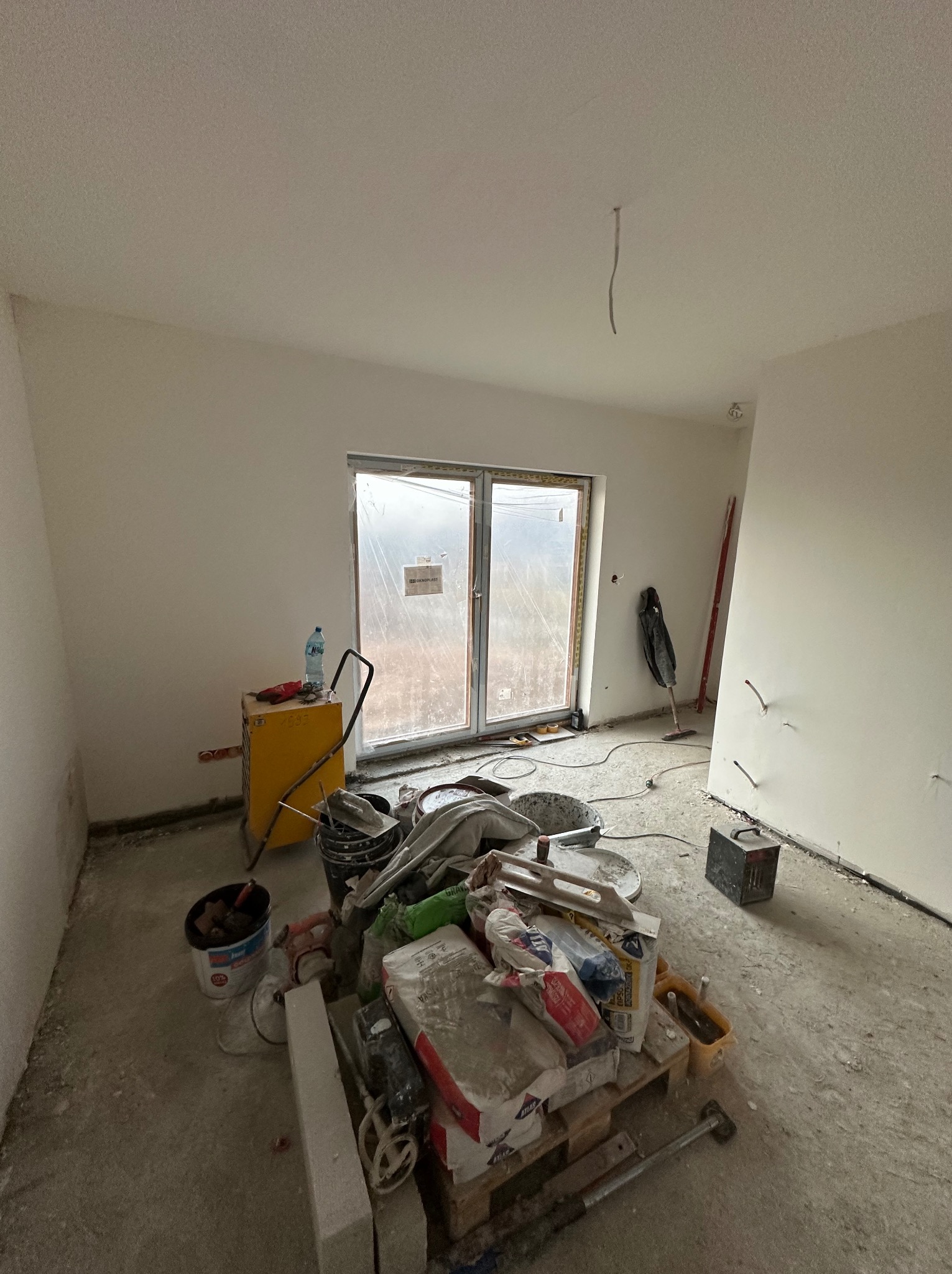 Wnętrze pokoju w trakcie prac wykończeniowych: świeżo pomalowane ściany, okno zabezpieczone folią, narzędzia i materiały budowlane na palecie. Widoczne ślady prac na podłodze.