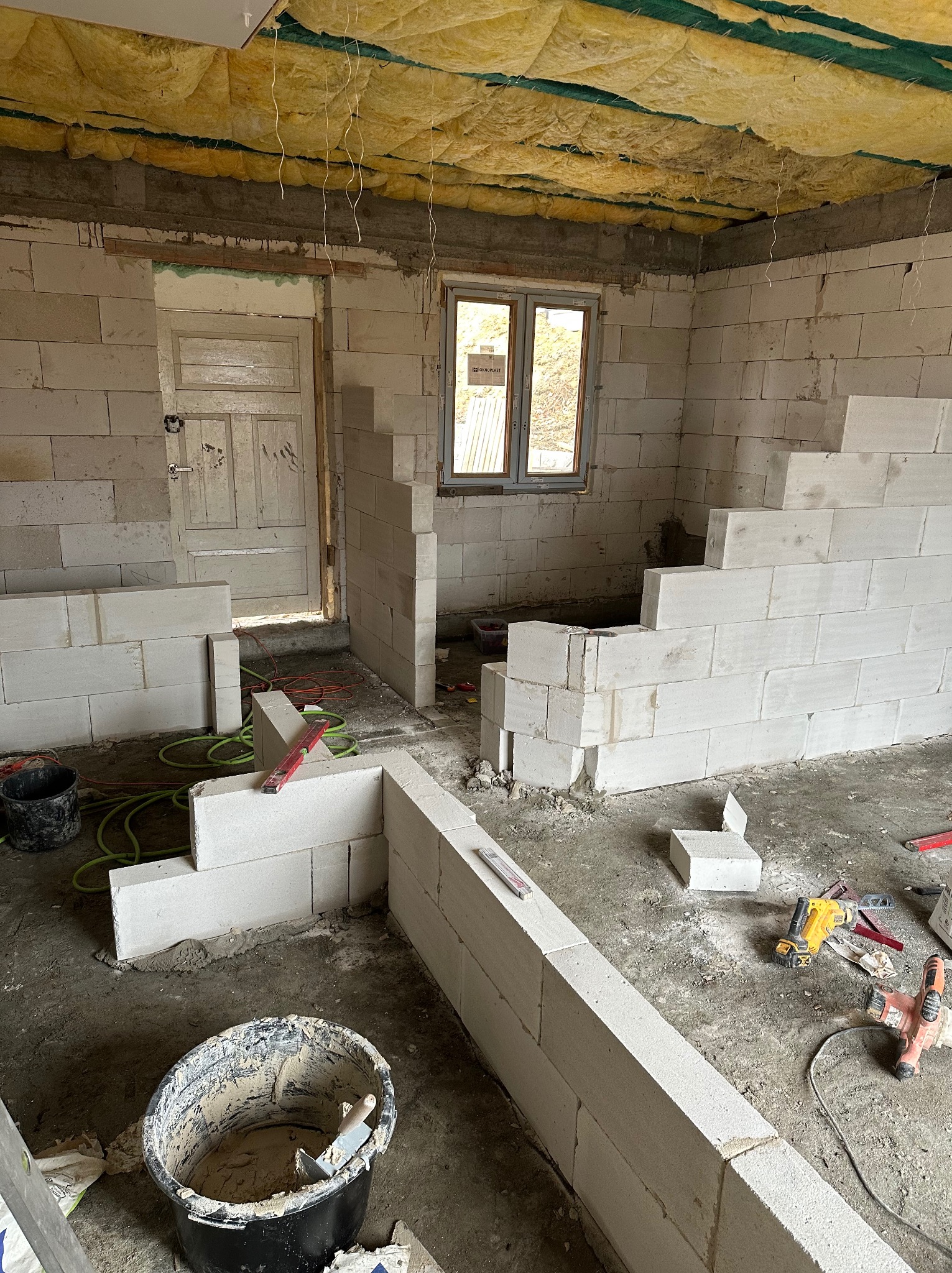 Wnętrze w trakcie murowania ścian z białych bloczków, widoczne narzędzia, wiadro z zaprawą i ocieplenie sufitu. Częściowo wymurowane ściany działowe.