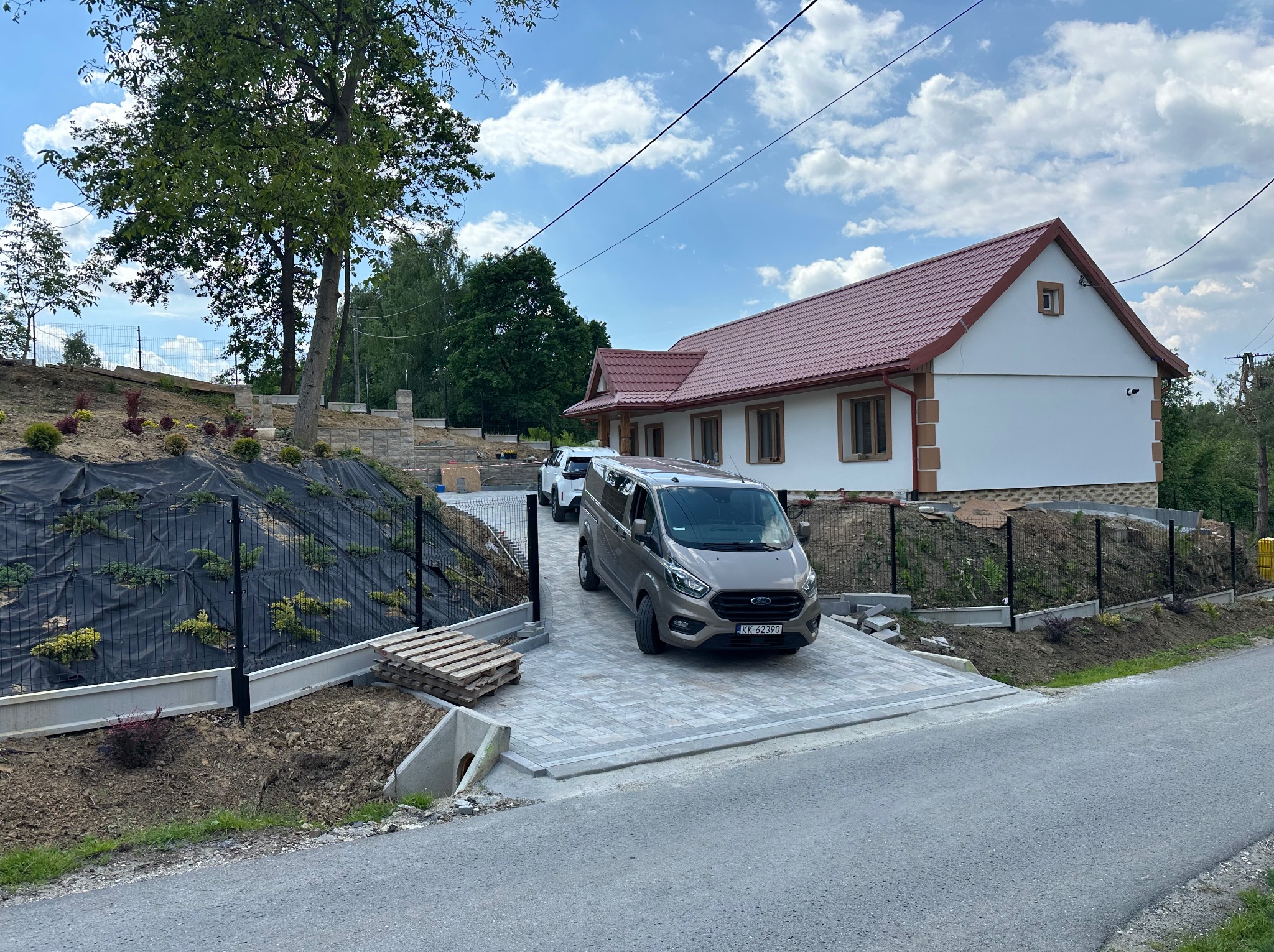 Nowo wybudowany dom z czerwonym dachem i beżowymi akcentami, widoczny z podjazdu wyłożonego kostką. Na podjeździe stoi szary van, a w tle widać drugi samochód.